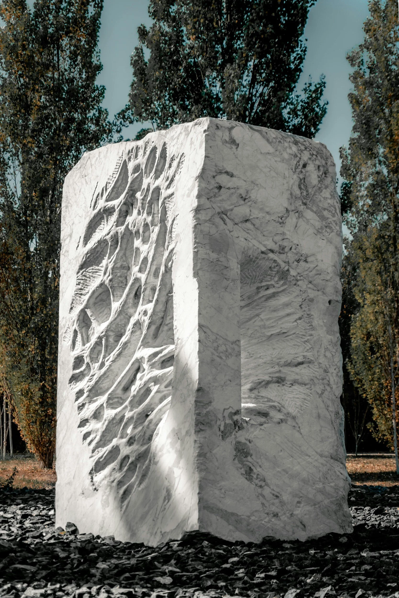 Skulptur aus weißem Marmor mit groben und feinen Maserungen, steil stehendes Block auf einem dunklen Kiesbett, umgeben von Bäumen mit grünen Blättern, bei Tageslicht.