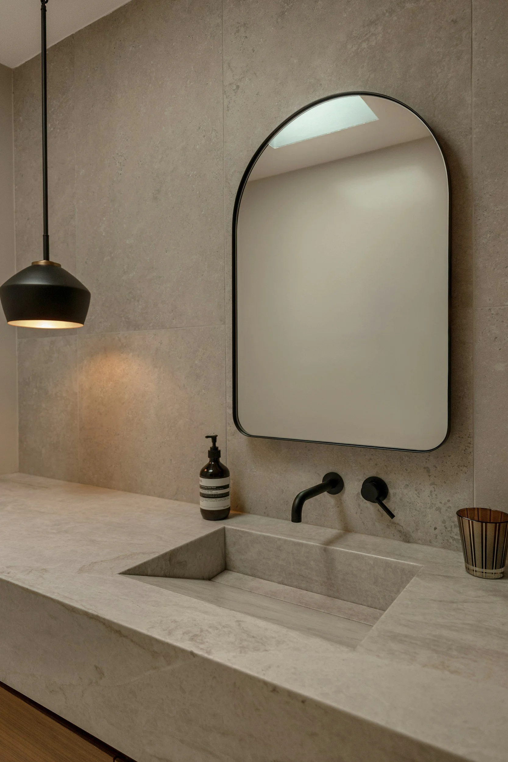 Moderner Badezimmerwaschplatz mit einem schwarzen Armaturen, einem beige Steinwaschbecken, Spiegel, einer schwarzen Lampe, einer Seifenspender und einem kleinen Korb an einer gepflasterten Wand.