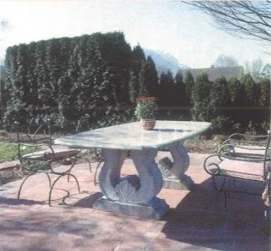 Ein runder Steintisch mit einer Pflanze in einer Blumenvase in der Mitte, umgeben von Metallstühlen auf einer Terrasse mit Garten im Hintergrund.