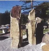 Drei große aufrechte Steine auf einem Rasen, im Hintergrund Bäume und Autos.