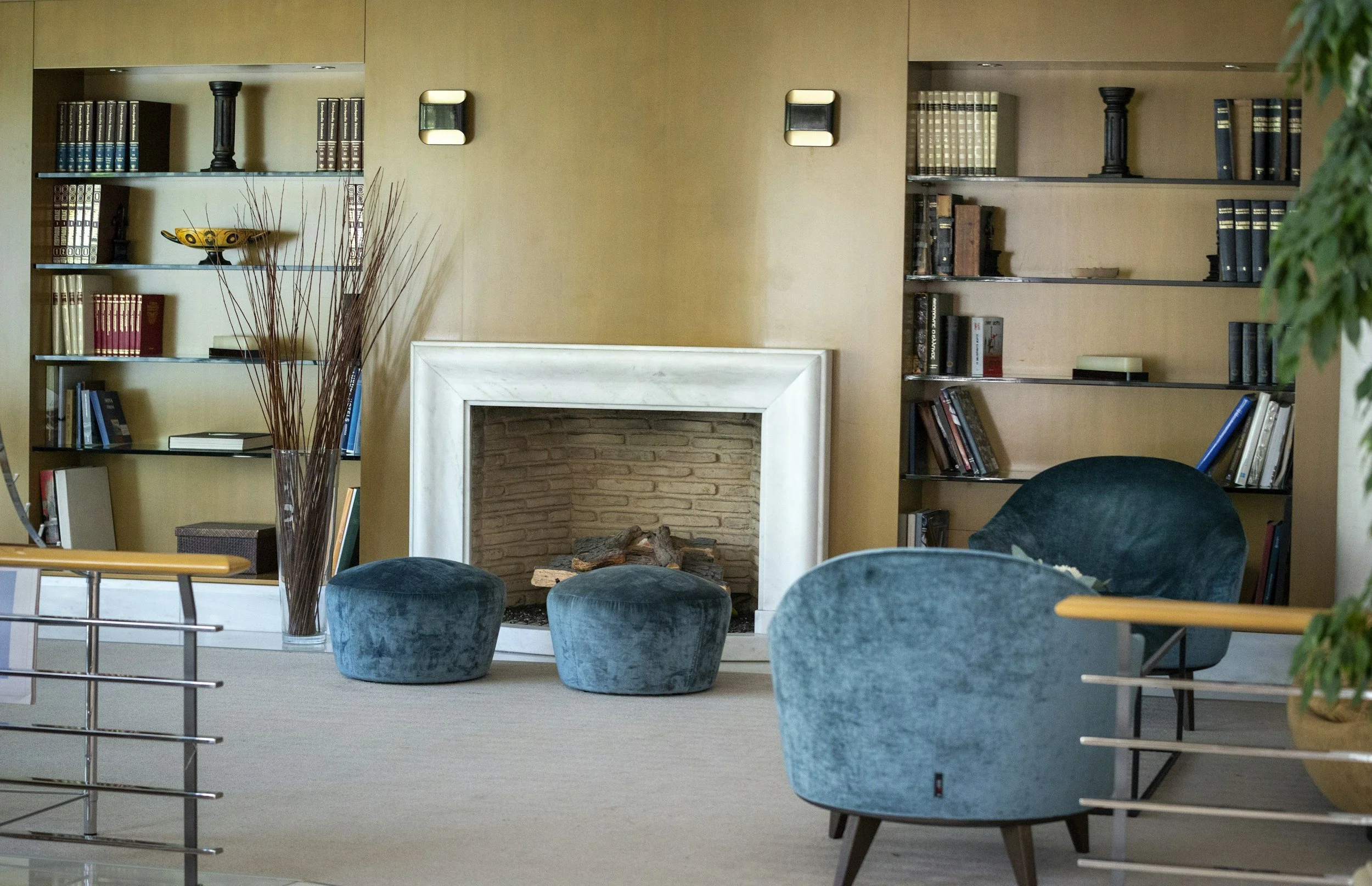 Ein gemütliches Wohnzimmer mit einem Kamin, Bücherregalen, einem blauen Sessel, einem Hocker und Dekorationen auf den Regalen.