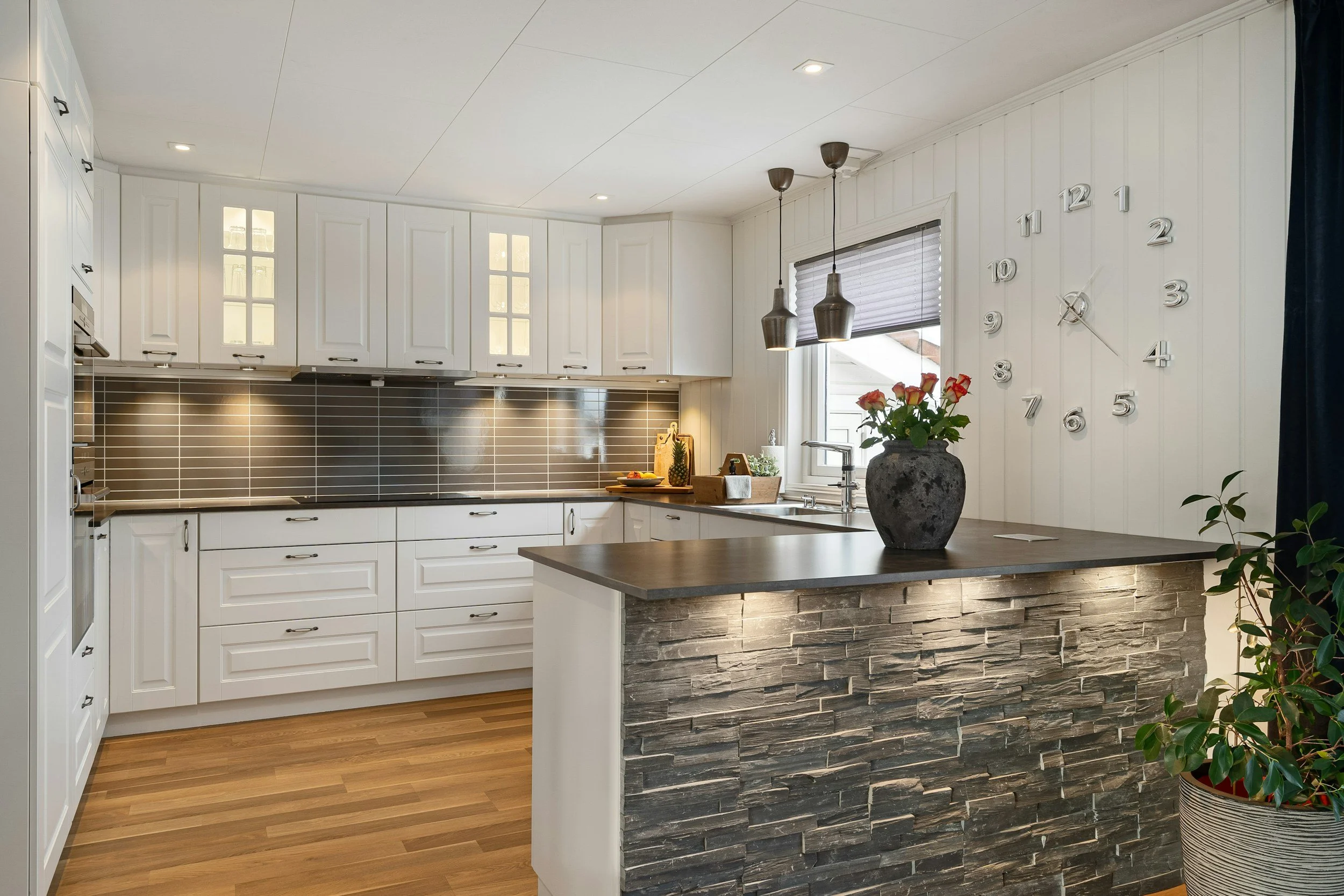 Moderne weiße Küche mit dunklem Backstein-Serviertresen, Topfpflanze auf Theke, Küchenschränken, Wanduhr, Fenster, hängese Lampen, Holzboden.