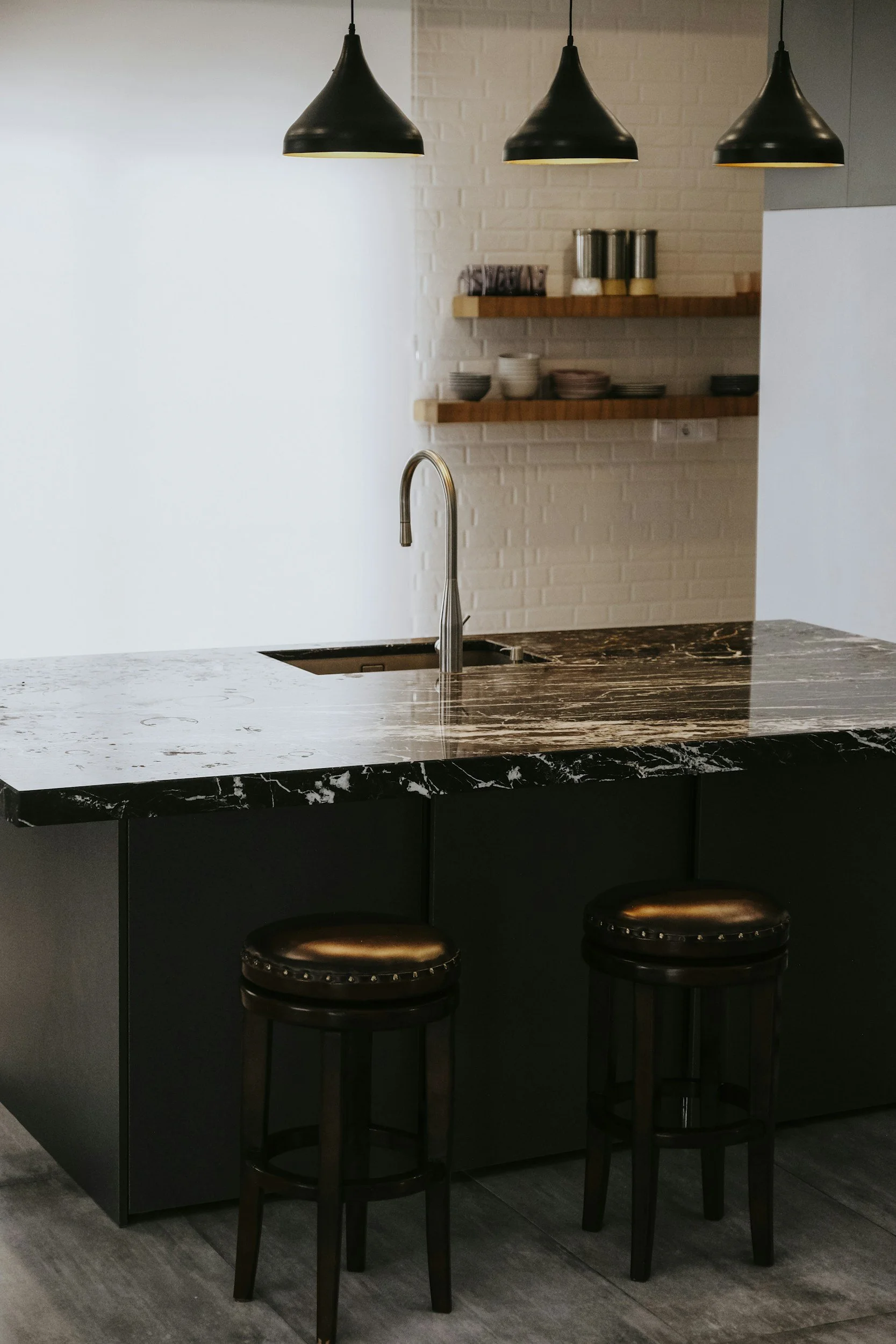 Moderne Küche mit schwarzen Barhockern, einer schwarzen Marmortheke, einem Wasserhahn, einer weißen Ziegelwand und offenen Holzregalen mit Geschirr und Gewürzen, beleuchtet von drei schwarzen Pendelleuchten.
