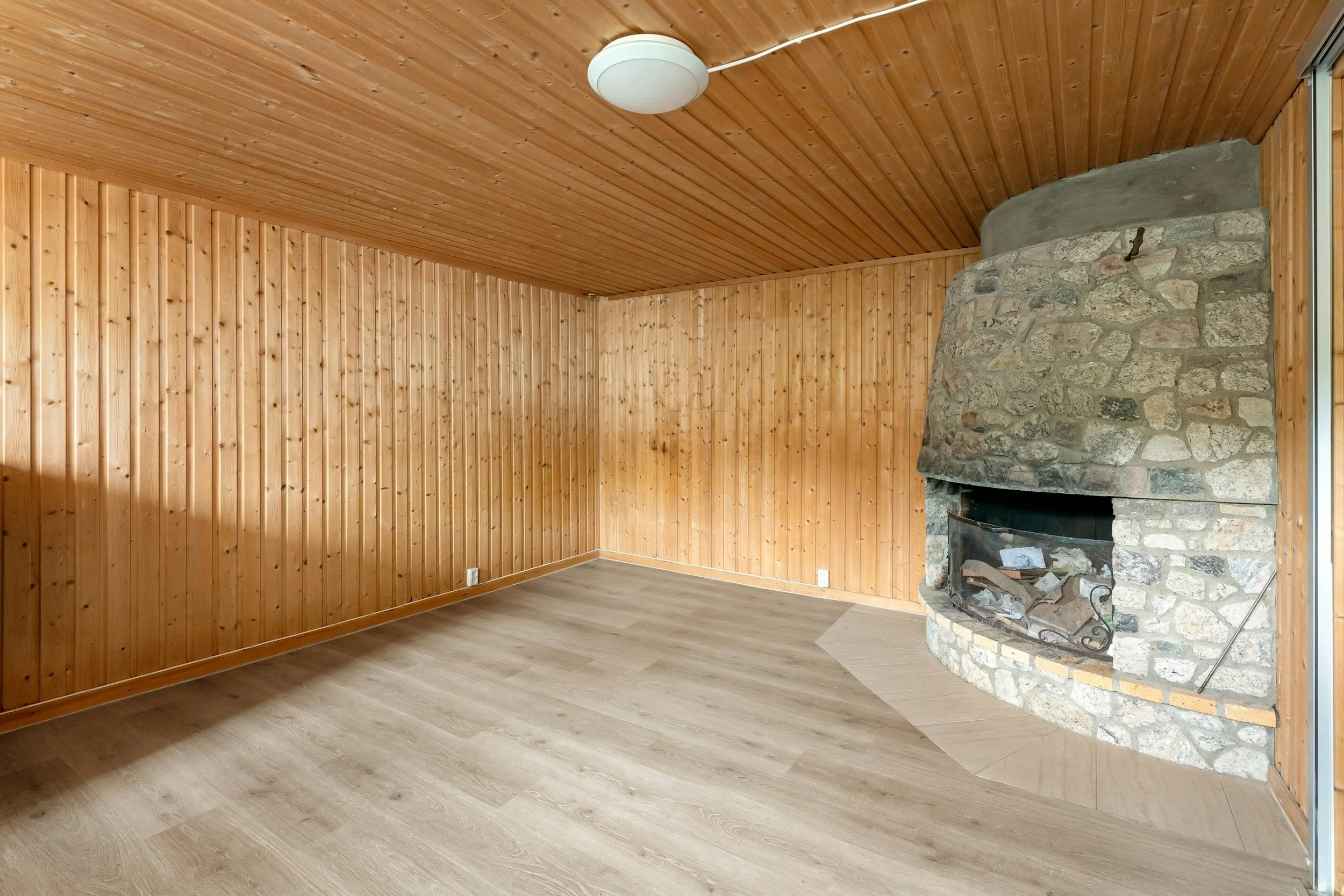 Innenraum mit Holzvertäfelung, Holzdielenboden und einem großen Steinkamin in der Ecke.