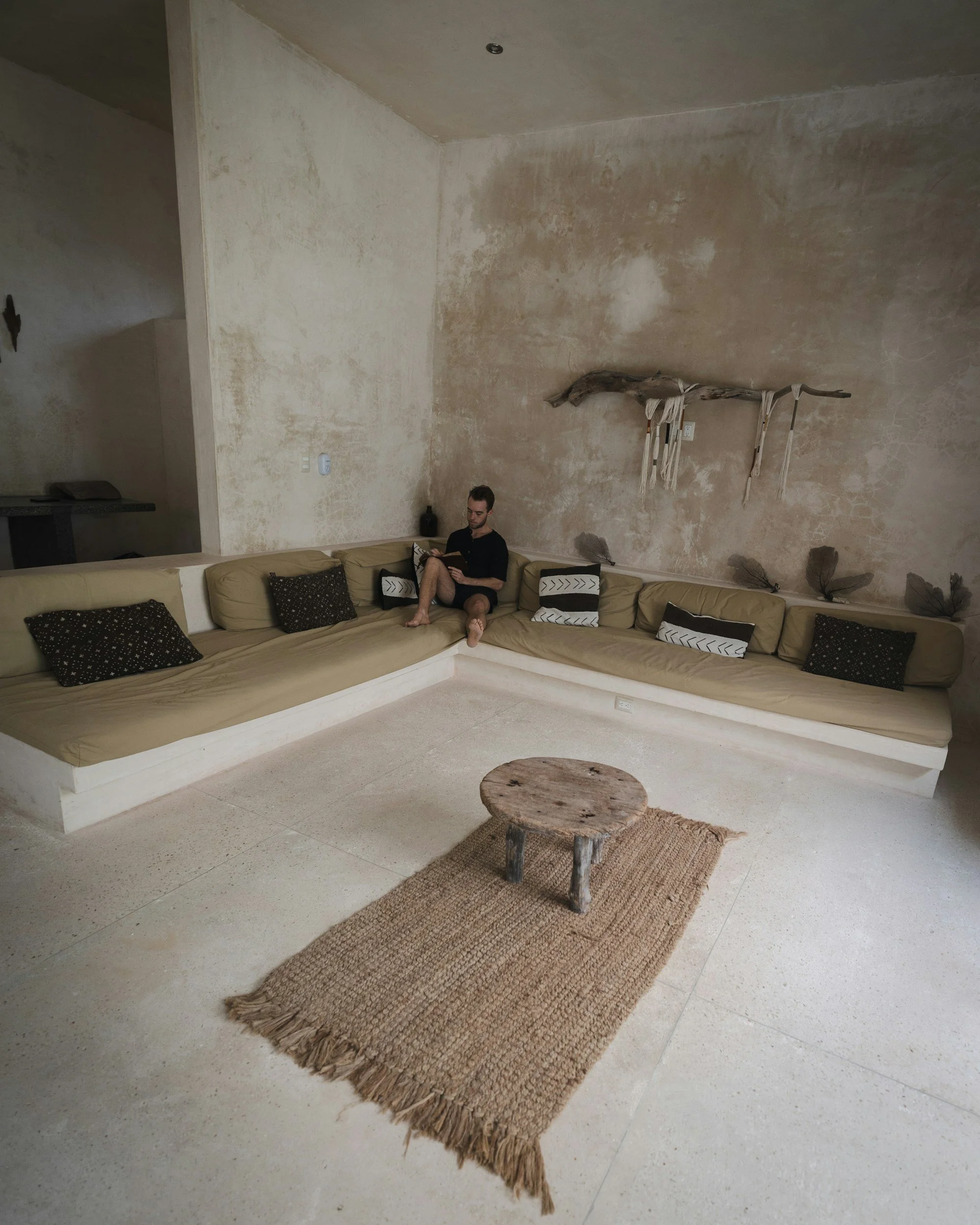 Wohnzimmer mit beige Couch, mehreren schwarzen und weißen Kissen, einem kleinen rustikalen Holztisch auf einem Juteteppich, einer Person sitzt auf der Couch und liest.