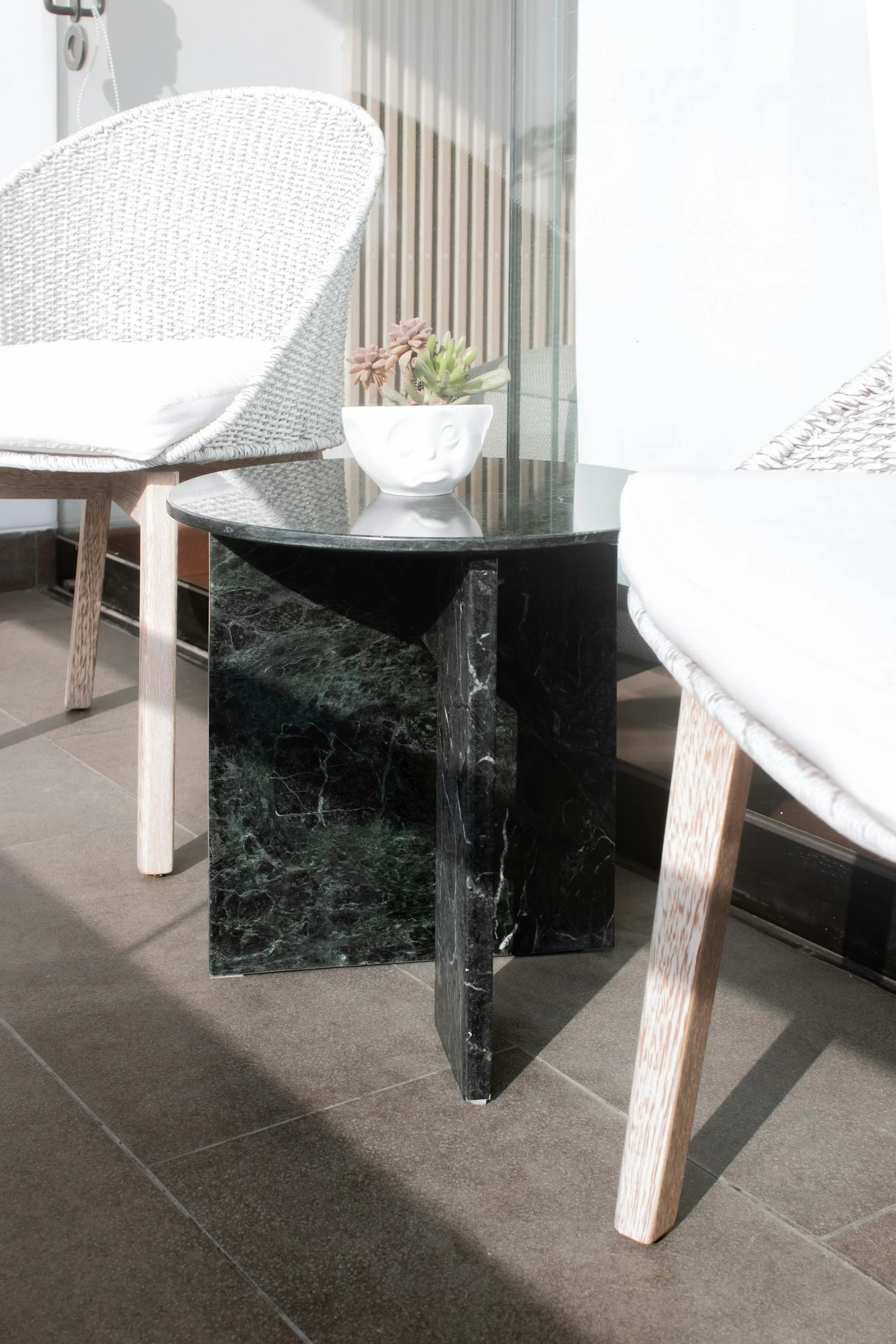 Ein schwarzer Marmor-Nachtisch zwischen zwei weißen Stühlen mit Holzbeinen, auf dem eine weiße Pflanzenschale mit Sukkulenten steht.