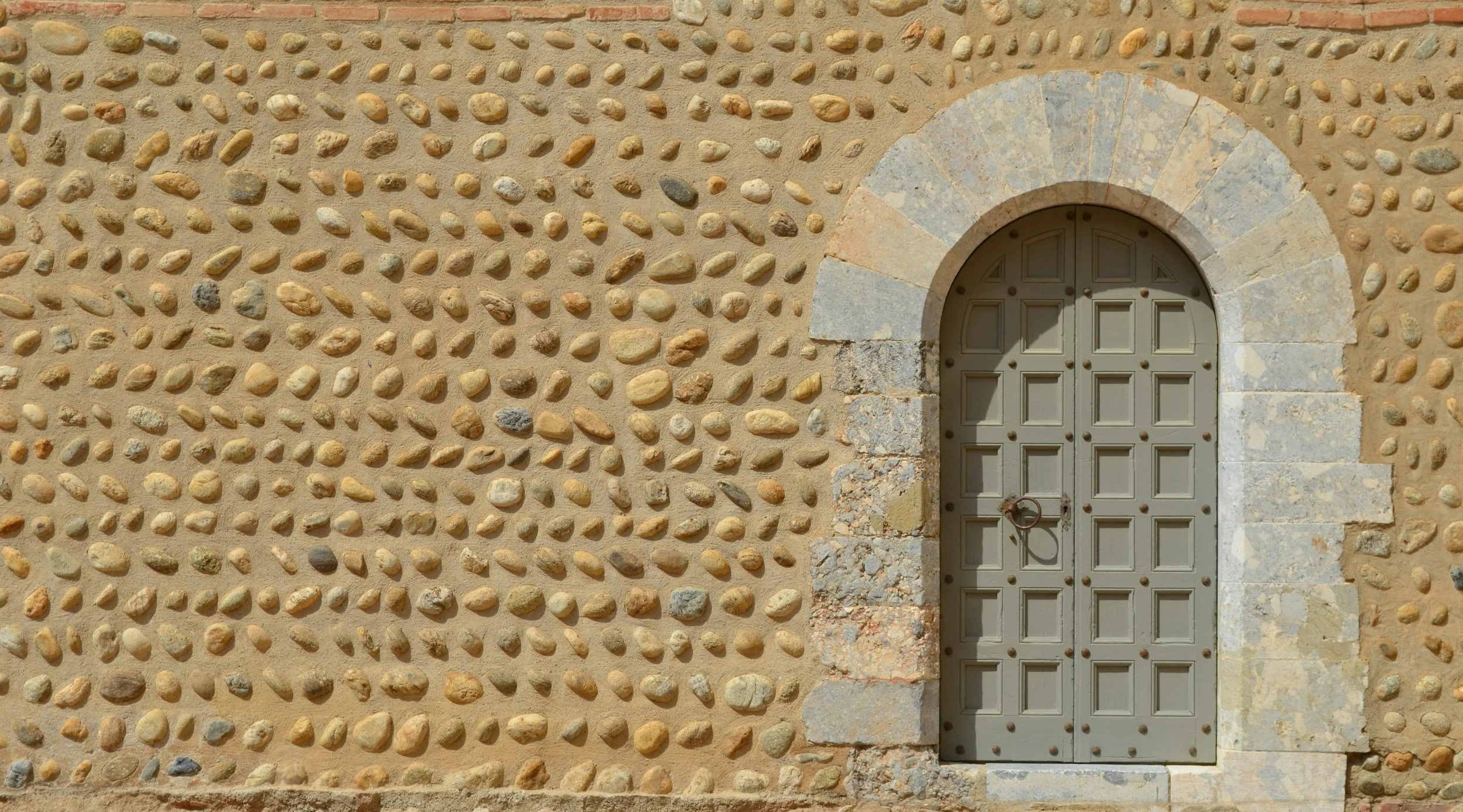 Ein Steinmauer mit rechteckigen und runden Steinen und einer runden Tür aus Holz mit Eisenbeschlägen und einem Türklopfer. Die Tür ist in einem Steinrahmen eingebettet.