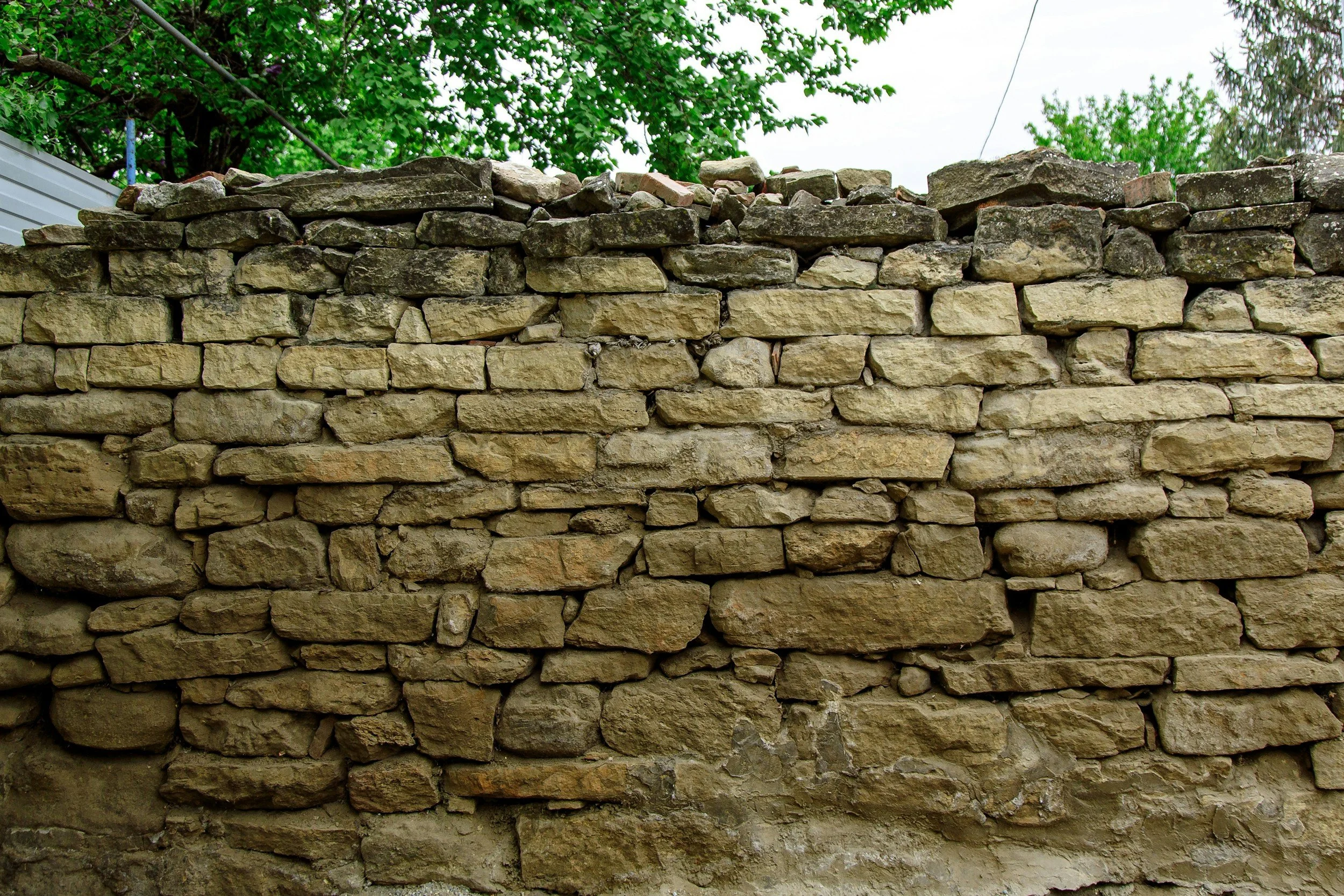 Ein Sichtblick auf eine alte, unregelmäßig gebaute Steinmauer mit grünen Bäumen im Hintergrund.