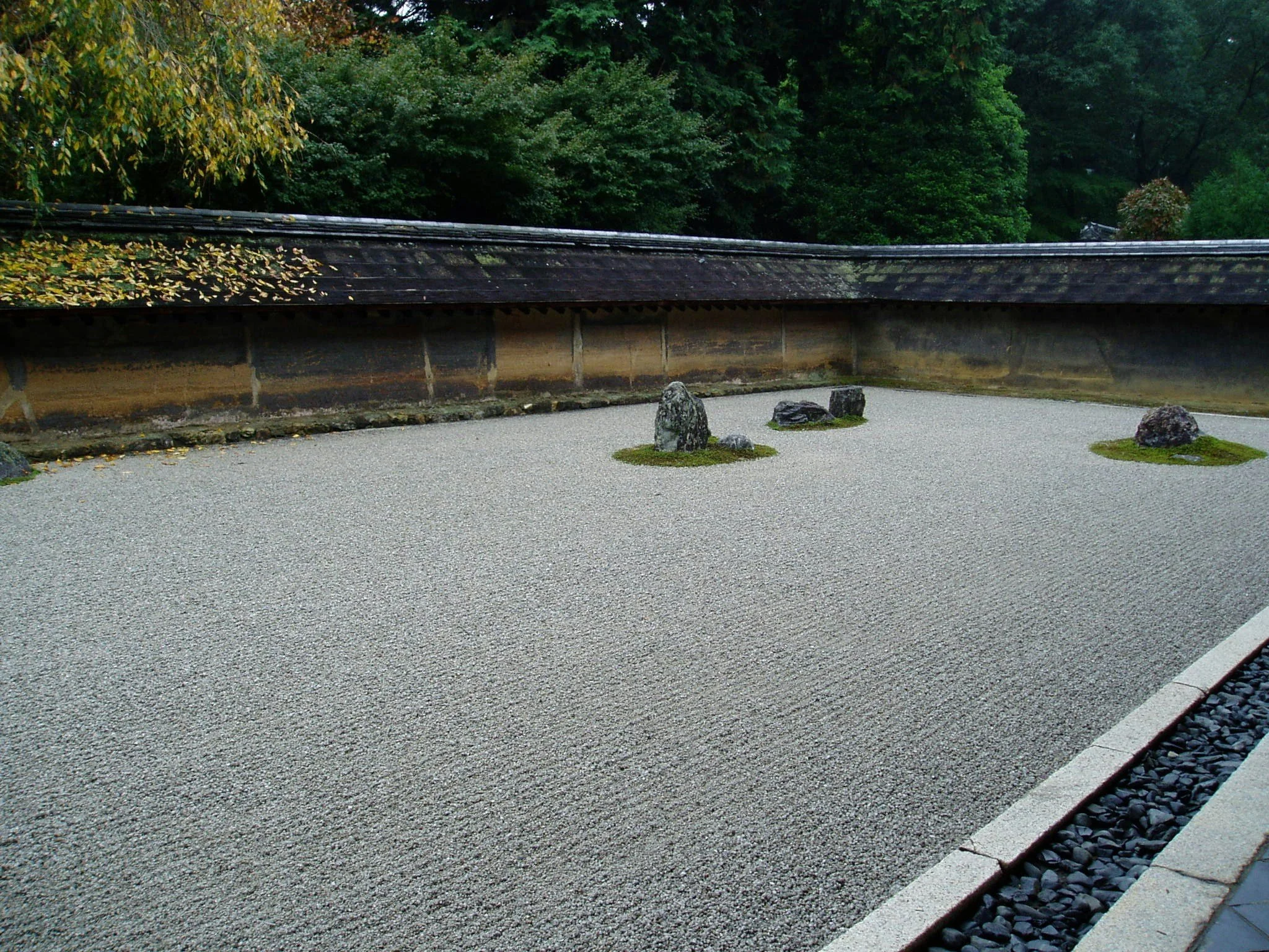 Ein japanischer Zen-Garten mit Kies, Steinen und Moos, eingezäunt von einer holzvertäfelten Mauer und Bäumen im Hintergrund.