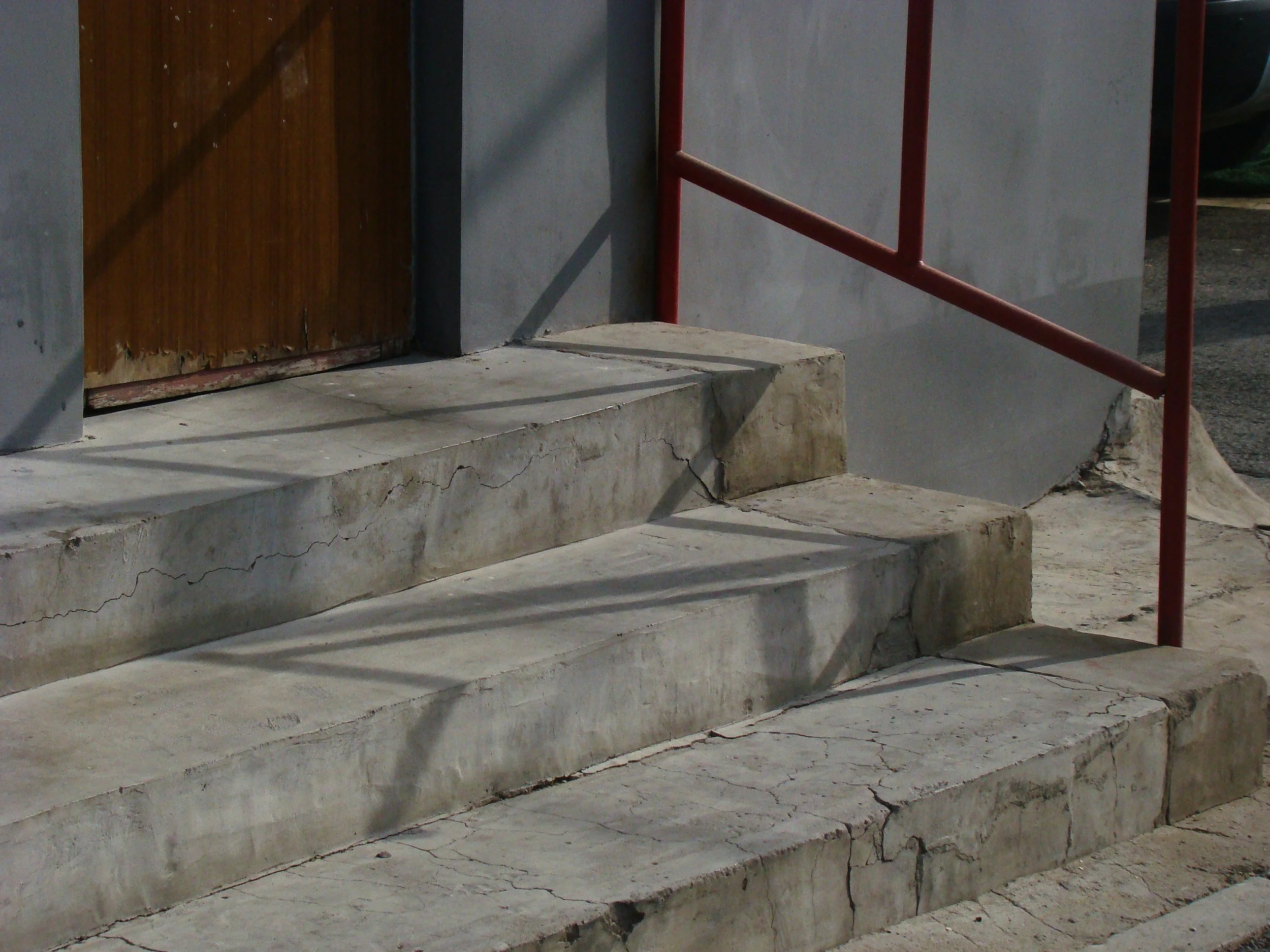 Alte, beschädigte Betonstufen vor einer Tür mit Holztür und roter Metallhandlauf