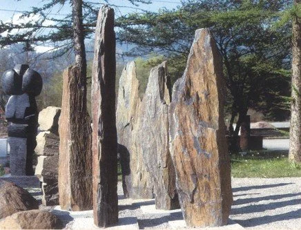 Mehrere große, vertikale Steine unterschiedlicher Größen und Formen in einem Freiluft-Garten, umgeben von Bäumen und Schatten.
