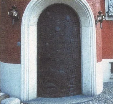 Schwarze Tür mit kreisförmigen Mustern, umgeben von einem weißen Steinrahmen, an einem roten Gebäude mit Laternen