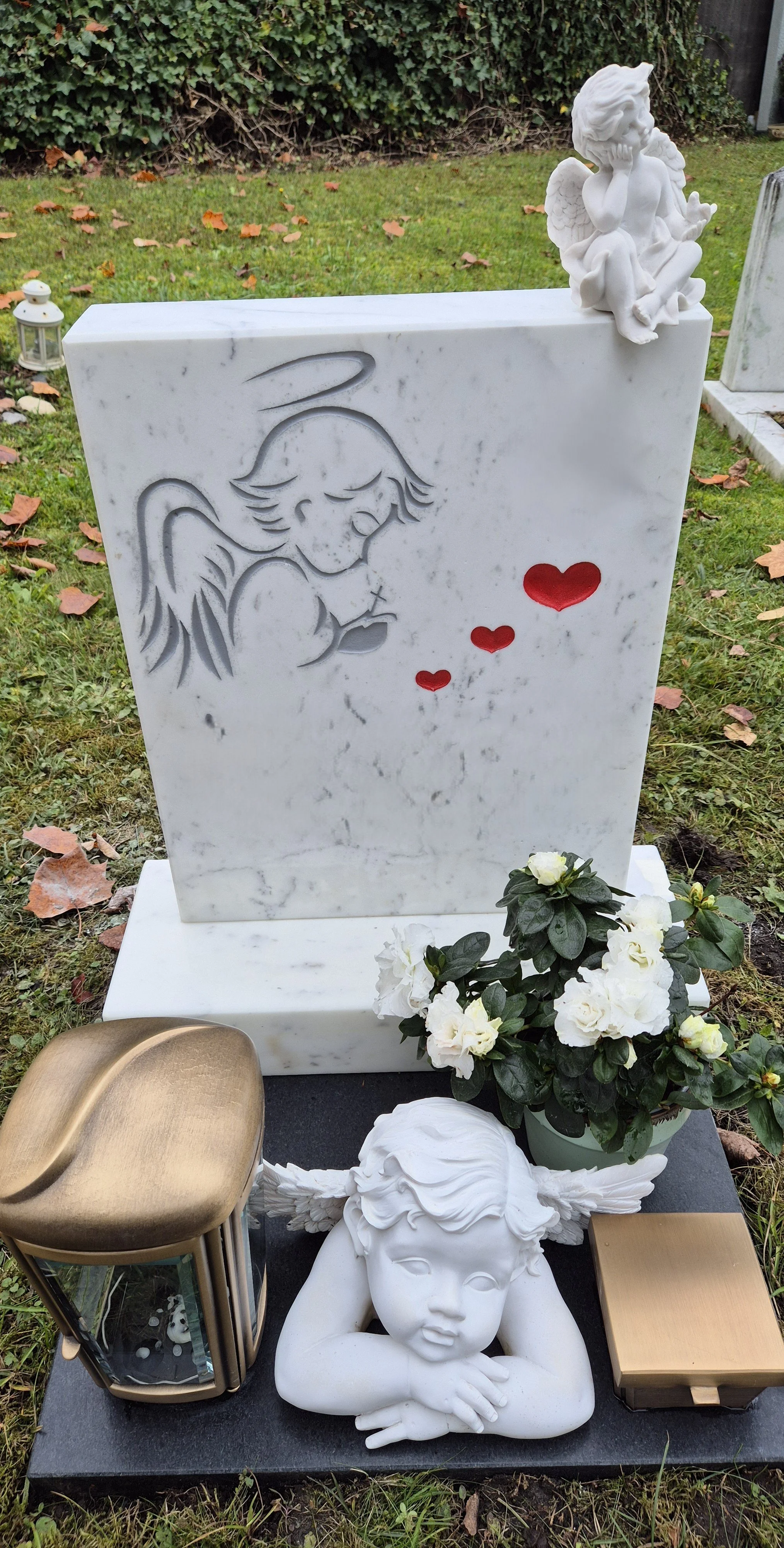 Ein Grabstein mit einer eingravierten Zeichnung eines Komas. Auf dem Grabstein befindet sich eine kleine, weiße Engelsfigur und eine weiße Blume. Vor dem Grabstein liegen ein bronze- und ein goldfarbener Kerzenleuchter, eine weiße Figur eines Babys m