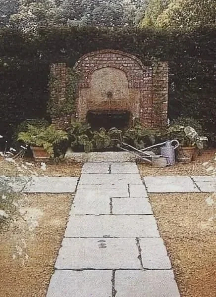 Ein Garten mit einem Kiesweg, Wasserhahn, Eimer und einem Steinbrunnen im Hintergrund, umgeben von Büschen.