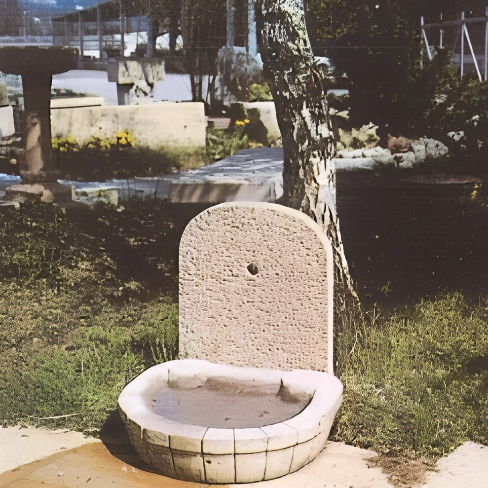 Eine Steinbank im Freien, umgeben von Büschen und einem Baum, im Hintergrund ein Weg und eine Mauer.