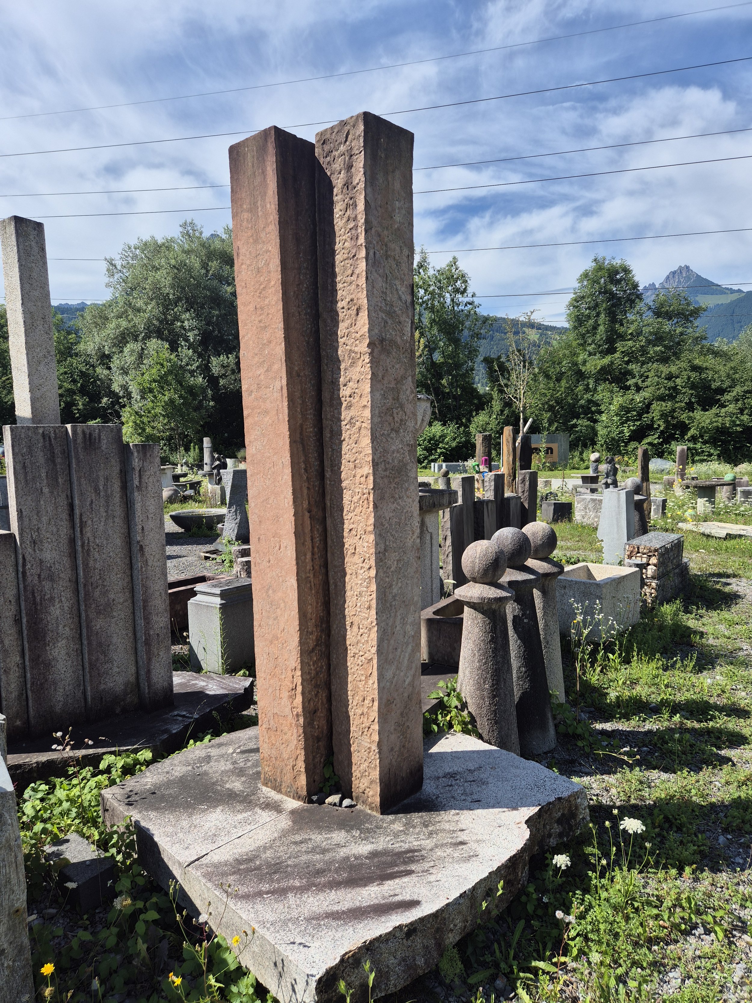Zwei große, vertikale Steinsäulen auf einem alten Friedhof, umgeben von weiteren Grabsteinen und Skulpturen, mit Bäumen und Bergen im Hintergrund.