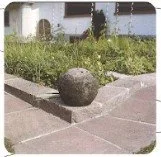 Ein großer runder Stein liegt auf einem gepflasterten Weg neben Gras und Pflanzen, mit einem Zaun im Hintergrund.