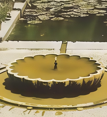 Gelber, blumenförmiger Wasserbrunnen mit einer Wasserquelle in der Mitte, im Außenbereich mit Wasserpflanzen im Hintergrund.