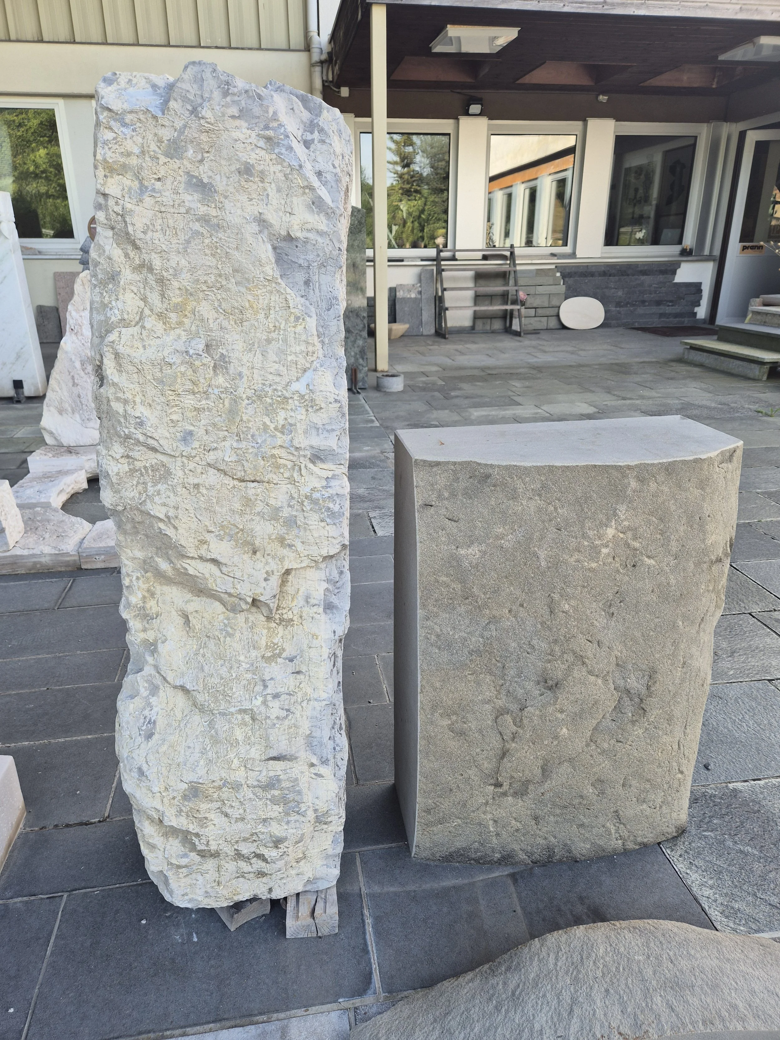 Zwei große Steinblöcke, einer mit weißlich-grauer Textur und der andere grau, stehen auf einer gepflasterten Terrasse vor einem Haus mit großen Fenstern und einer Holzüberdachung.