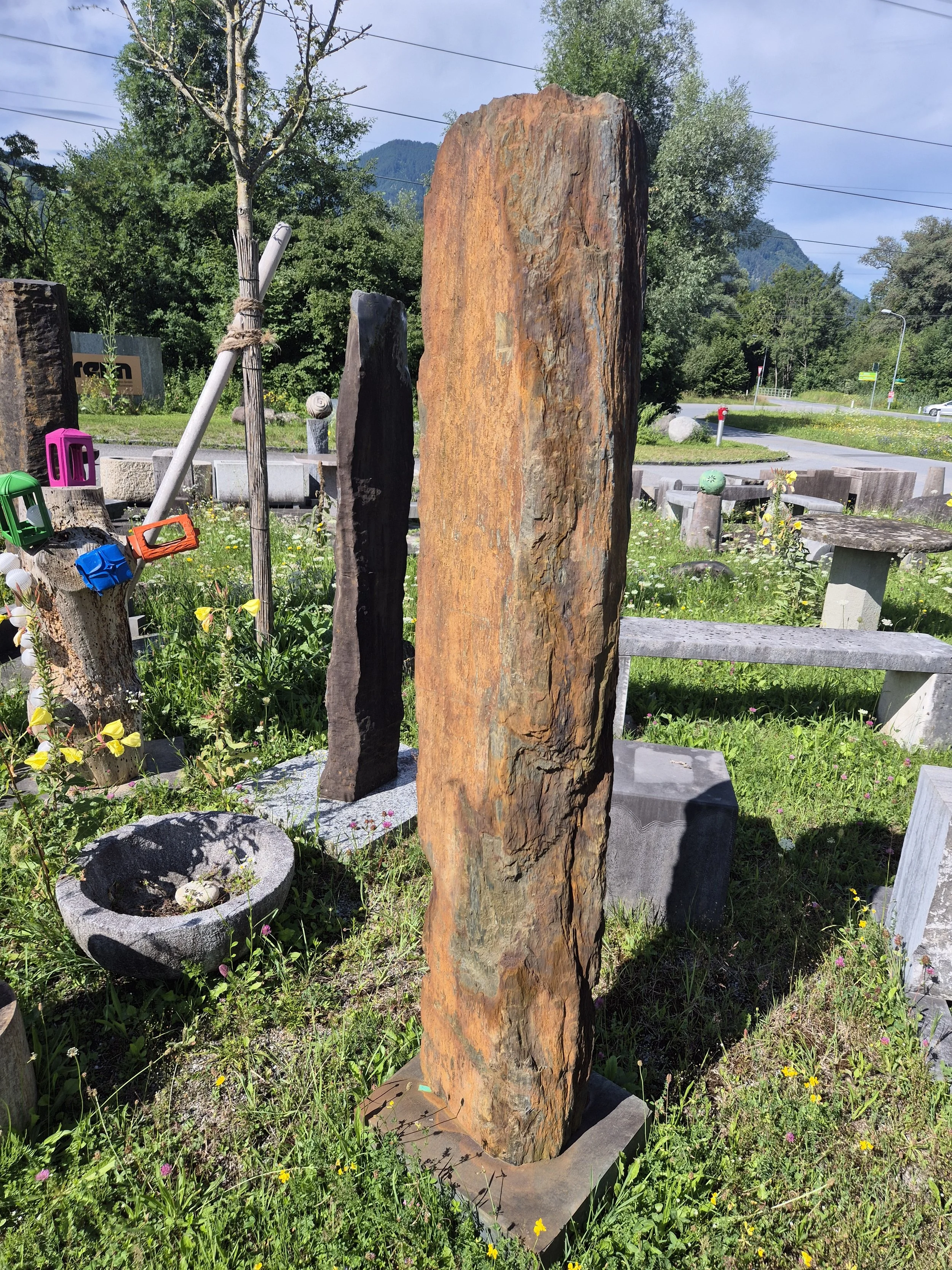 Mehrere große, rustikale Steinsäulen in einem Garten mit grünem Gras und bunten Blumen, im Hintergrund Bäume, eine Straße und Berge.