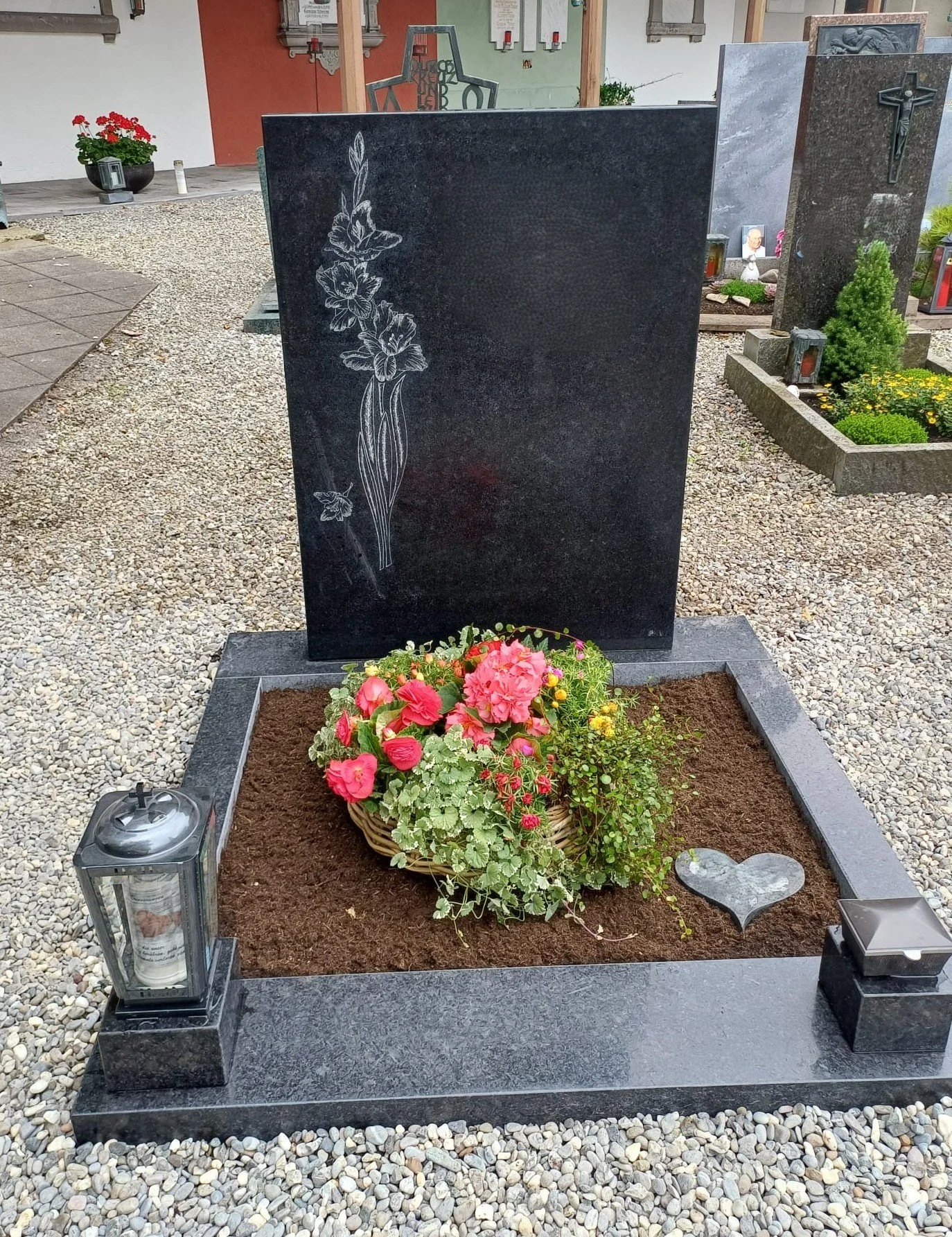 Grüner Grabstein mit Blumen und Kerzen im Friedhof, runde Blumenkomposition vor dem Stein, symbolischer Herzstein auf der Erde, umgeben von weiteren Grabsteinen und Pflanzen.