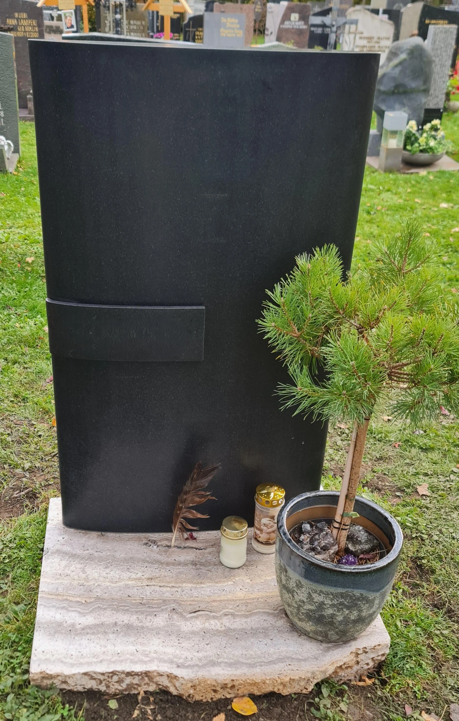 Ein schwarzer Grabstein auf einem Steinblock, eine kleine grüne Bonsai-Pflanze in einer Blumentopf, drei Kerzen in Gold- und Glasbehältern, und eine getrocknete Feder auf dem Steinblock, im Hintergrund ein Friedhof mit weiteren Gräbern und Grablten.