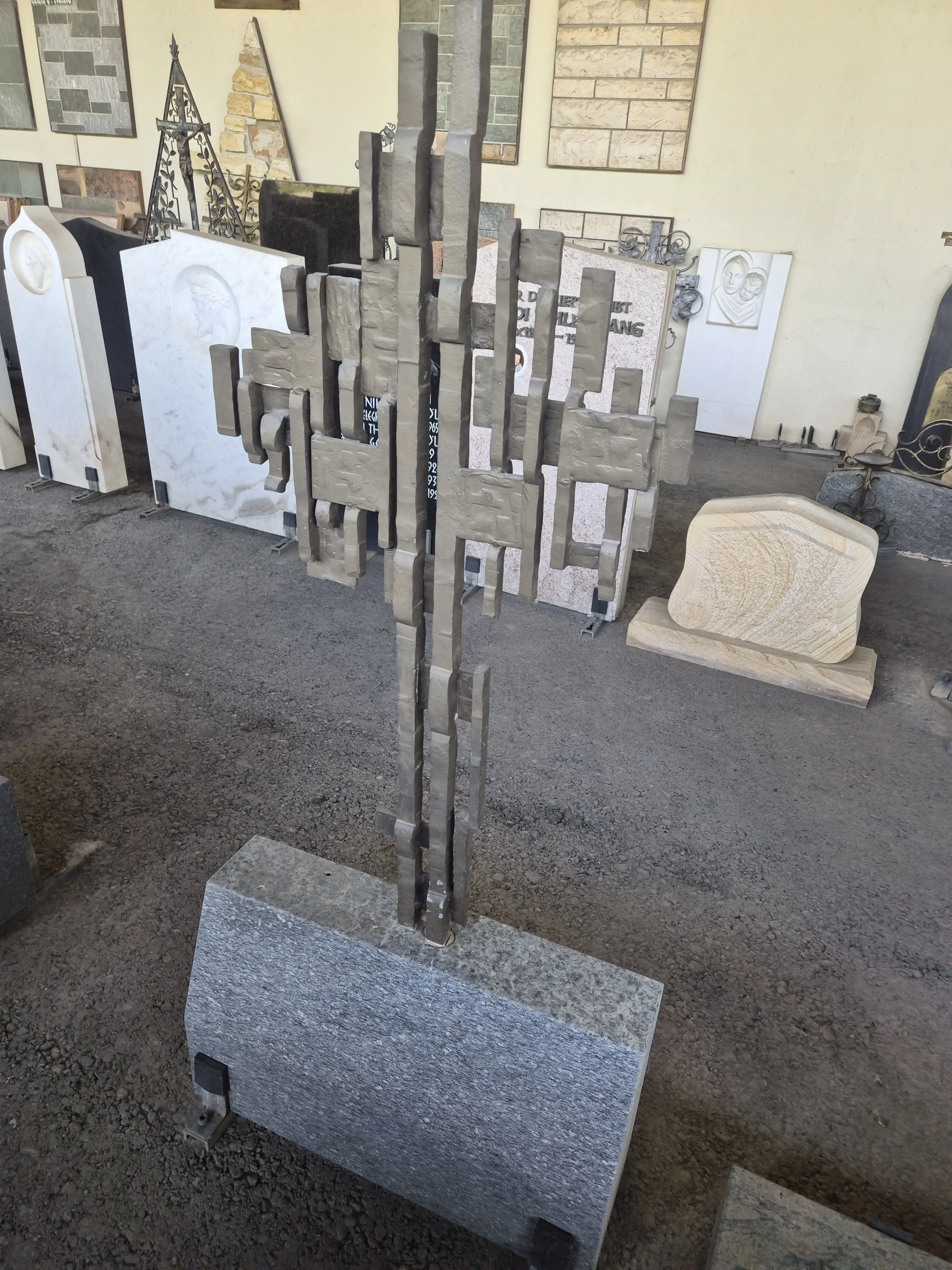 Künstlerische Skulptur aus Metall, die vertikal an einer quadratischen Granitbasis angebracht ist, in einem Ausstellungsraum mit verschiedenen Grabsteinen und Grabplatten im Hintergrund.