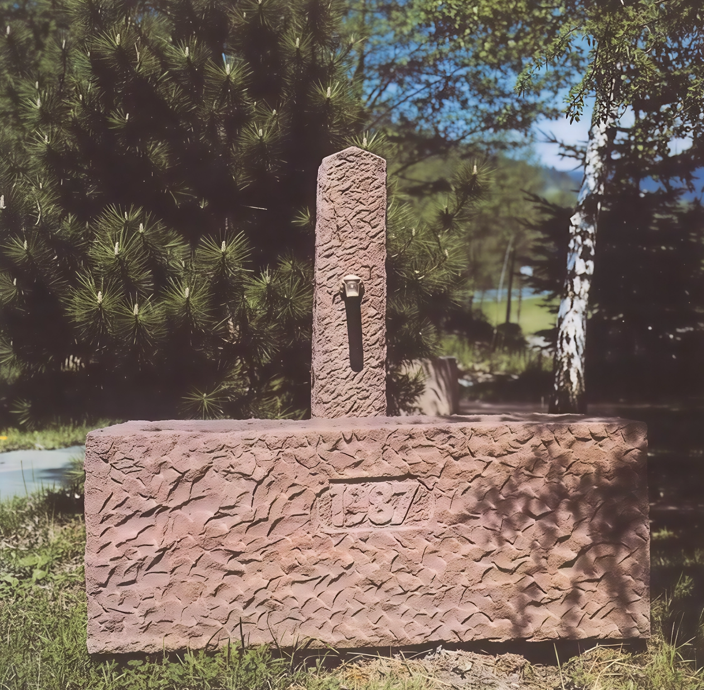 Eine Wasserstelle aus rotem, strukturiertem Stein im Freien mit einem Wasserhahn, umgeben von Bäumen und Natur.