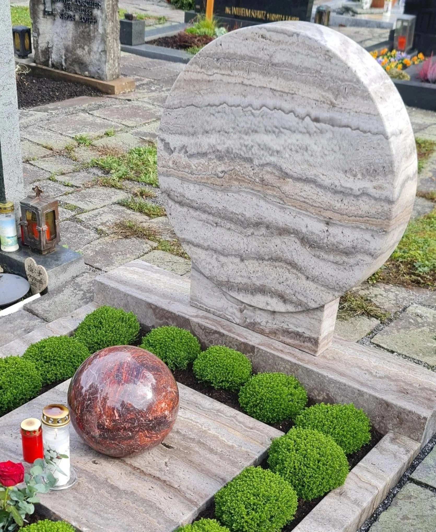 Grabhügel mit zwei Grabsteinen, einer großen aufrecht stehenden runden aus Marmor und einer kleineren kugelförmigen, roten Marmor-Statue. Es sind Gräber im Hintergrund sichtbar.