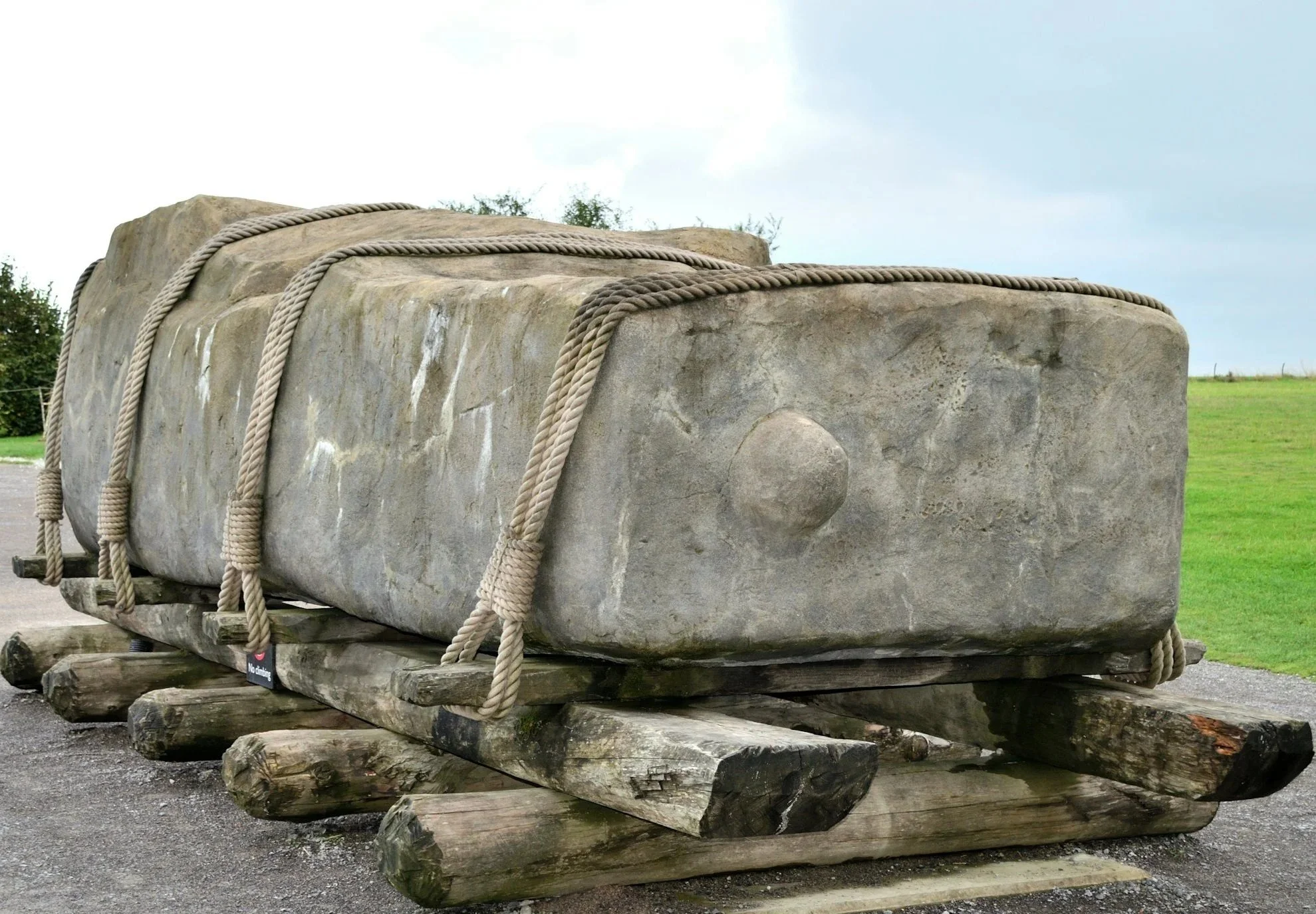 Großer prähistorischer Steinblock auf Holzplanken, mit Seilen gesichert, im Freien mit grüner Wiese im Hintergrund.