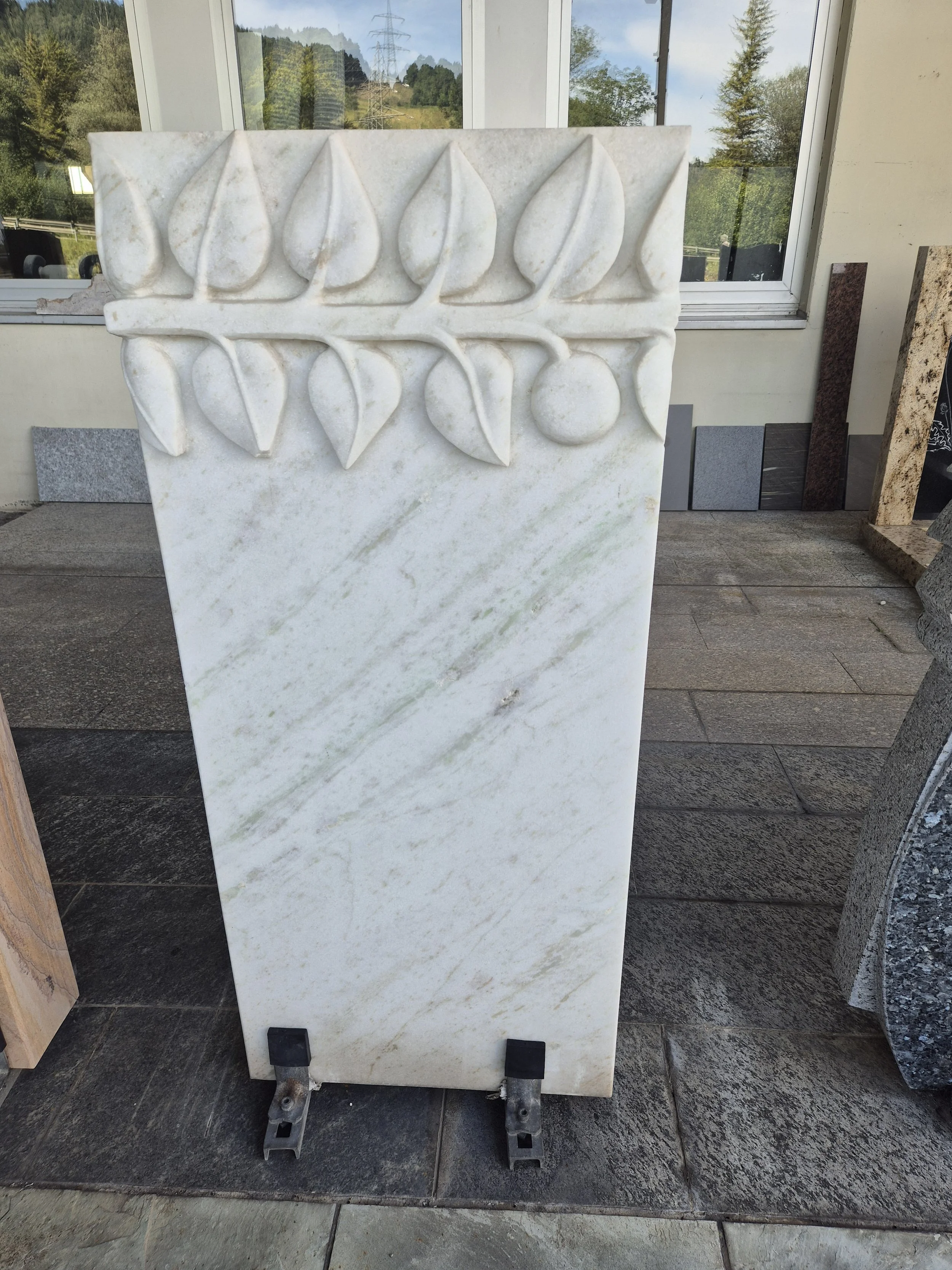 Ein weißes Marmor-Stein mit einer geschnitzten Verzierung von Blättern und einer Frucht am oberen Rand, stehender auf einem Betträger aus Metall, im Hintergrund Fenster mit Aussicht auf Natur und Stromleitungen.