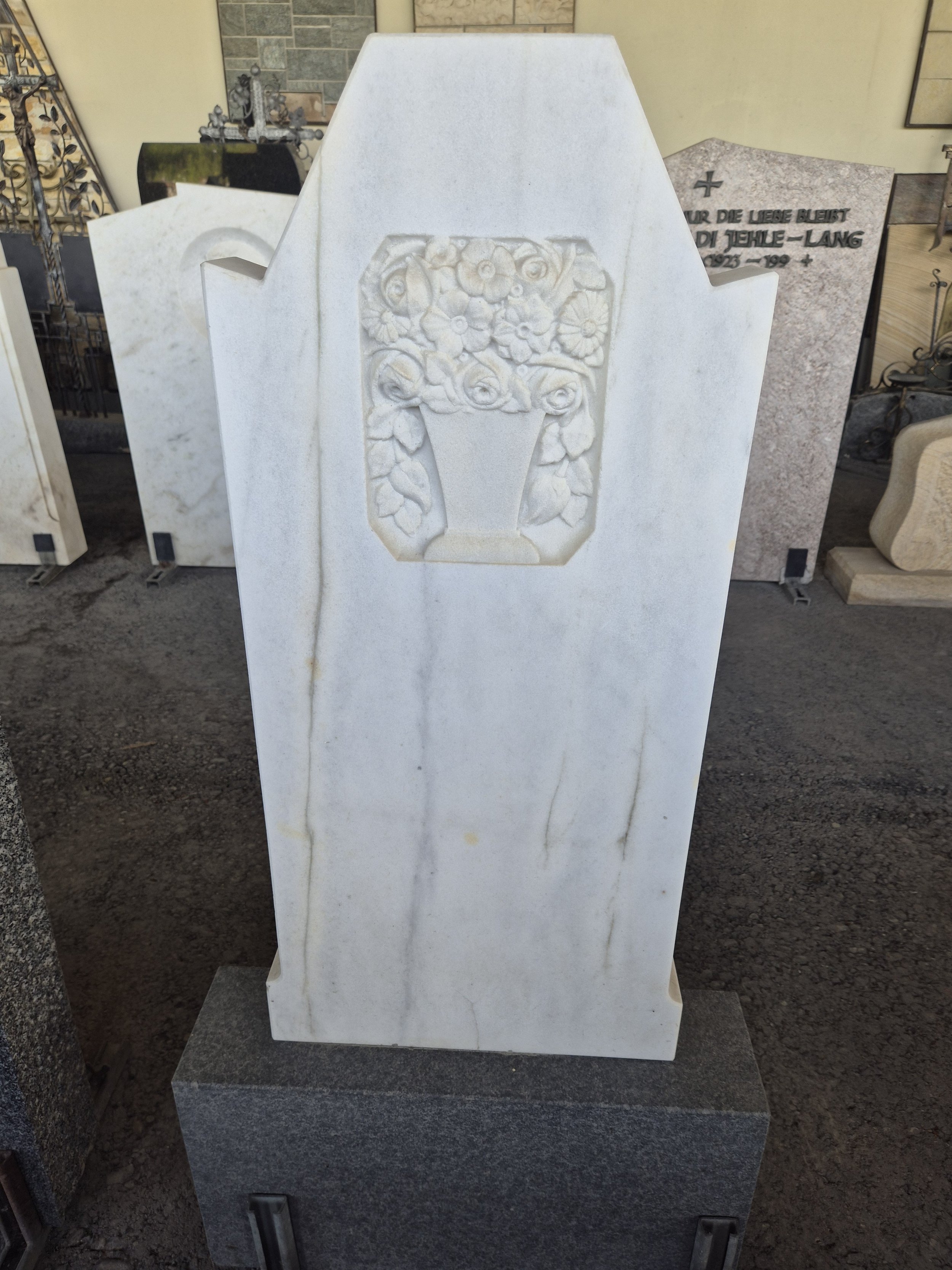 Grünes Grabstein mit Relief eines Blumenstraußes in einer Vase, auf einem weiteren Grabstein.