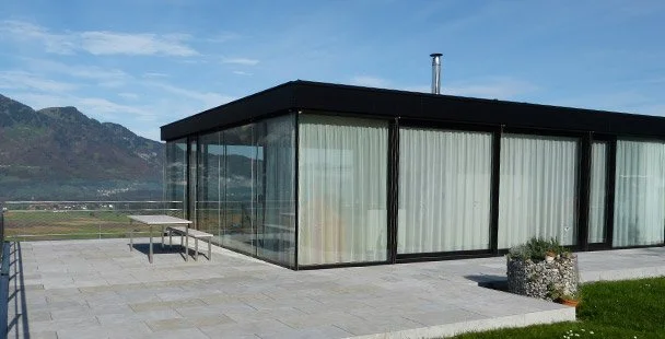 Modernes Gebäude mit großen Glasfenstern auf einer Terrasse mit Blick auf Berge.