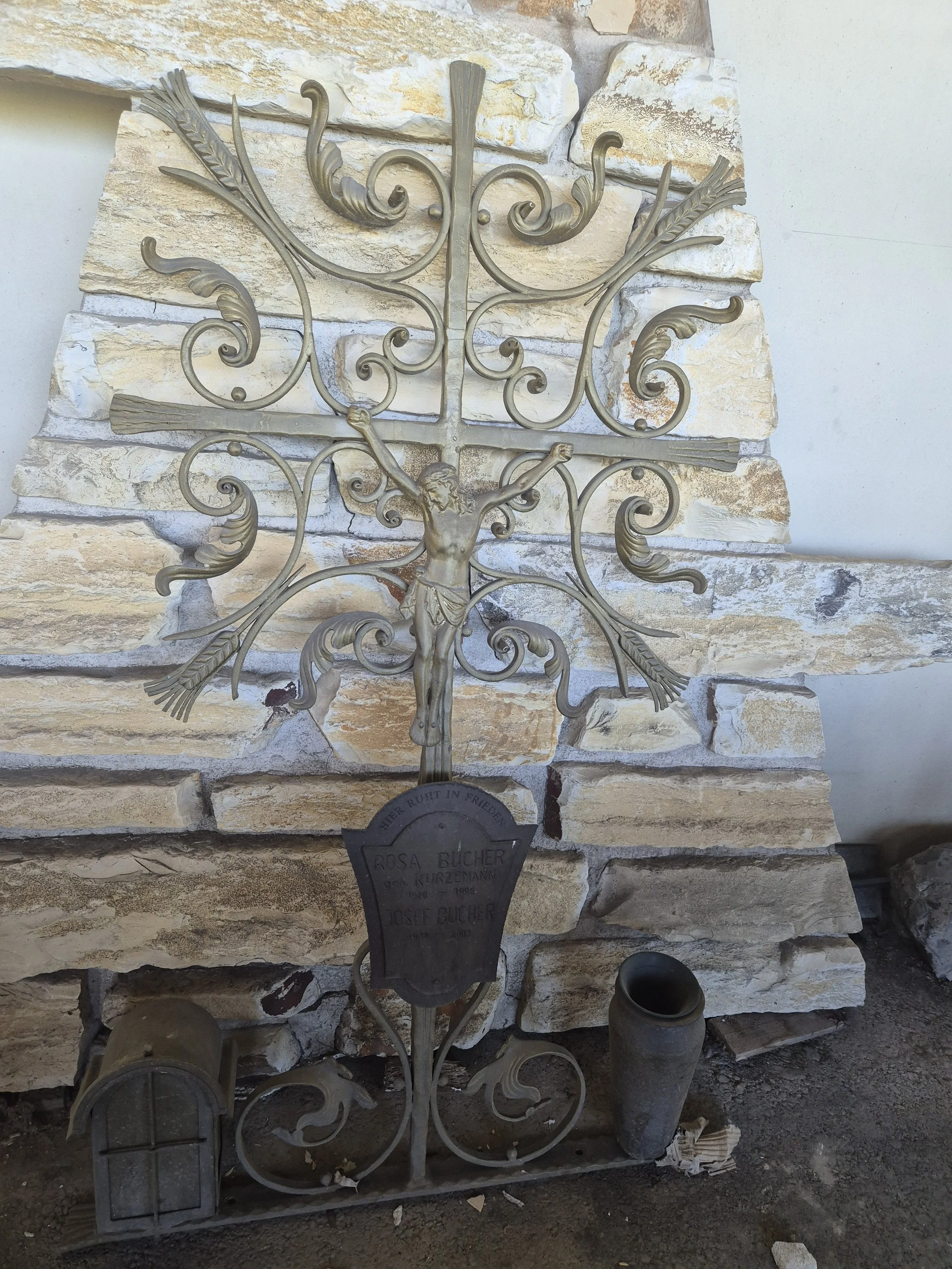 Metallkreuz mit Jesusfigur vor einem gemauerten Hintergrund, darunter ein schwarzer Grabstein mit der Aufschrift: "Hier ruht in Frieden Rosa Bucher geb. Kurzlehn 1910 - 1991, Josefa Bucher 1913".