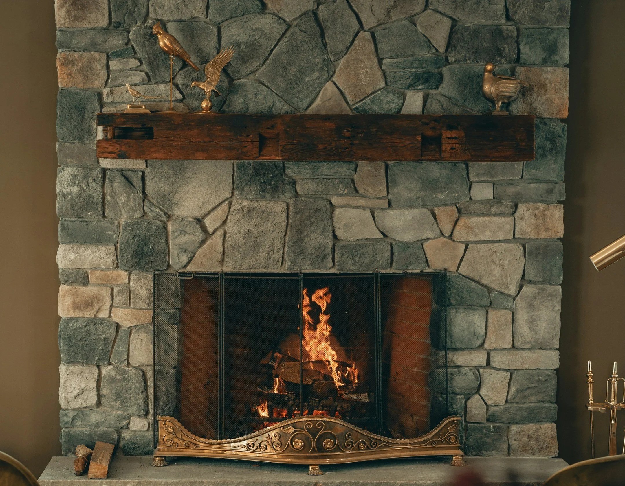 Ein Kamin aus unregelmäßig geformten Steinen mit brennendem Feuer, einer Holzablage mit goldfarbenen Skulpturen von Vögeln und einem goldfarbenen Kaminbildschirm.