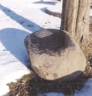 Ein großer, runder Stein mit einem geometrischen Muster auf der Oberseite, neben einem Baum und im Schnee.