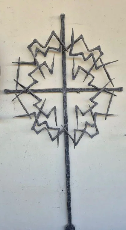 Ein kunstvolles Metall-Design, das wie ein Stern oder Sonnenrad aussieht, mit einem zentralen Kreuz und kreisförmigen Elementen, die an eine Sonne oder ein Rad erinnern.