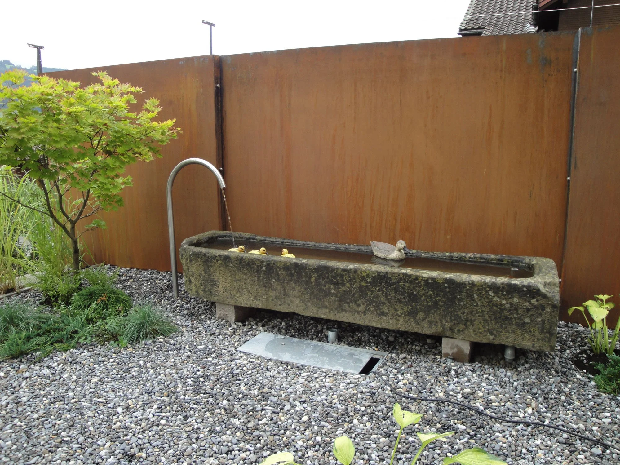 Ein alter Steintrog mit Wasser, eine Ente aus Kunststoff sitzt darin, daneben ein Wasserhahn, im Garten mit glasigem Kies als Boden, eine kleine grüne Pflanze auf der linken Seite und ein hoher Holzzaun im Hintergrund.