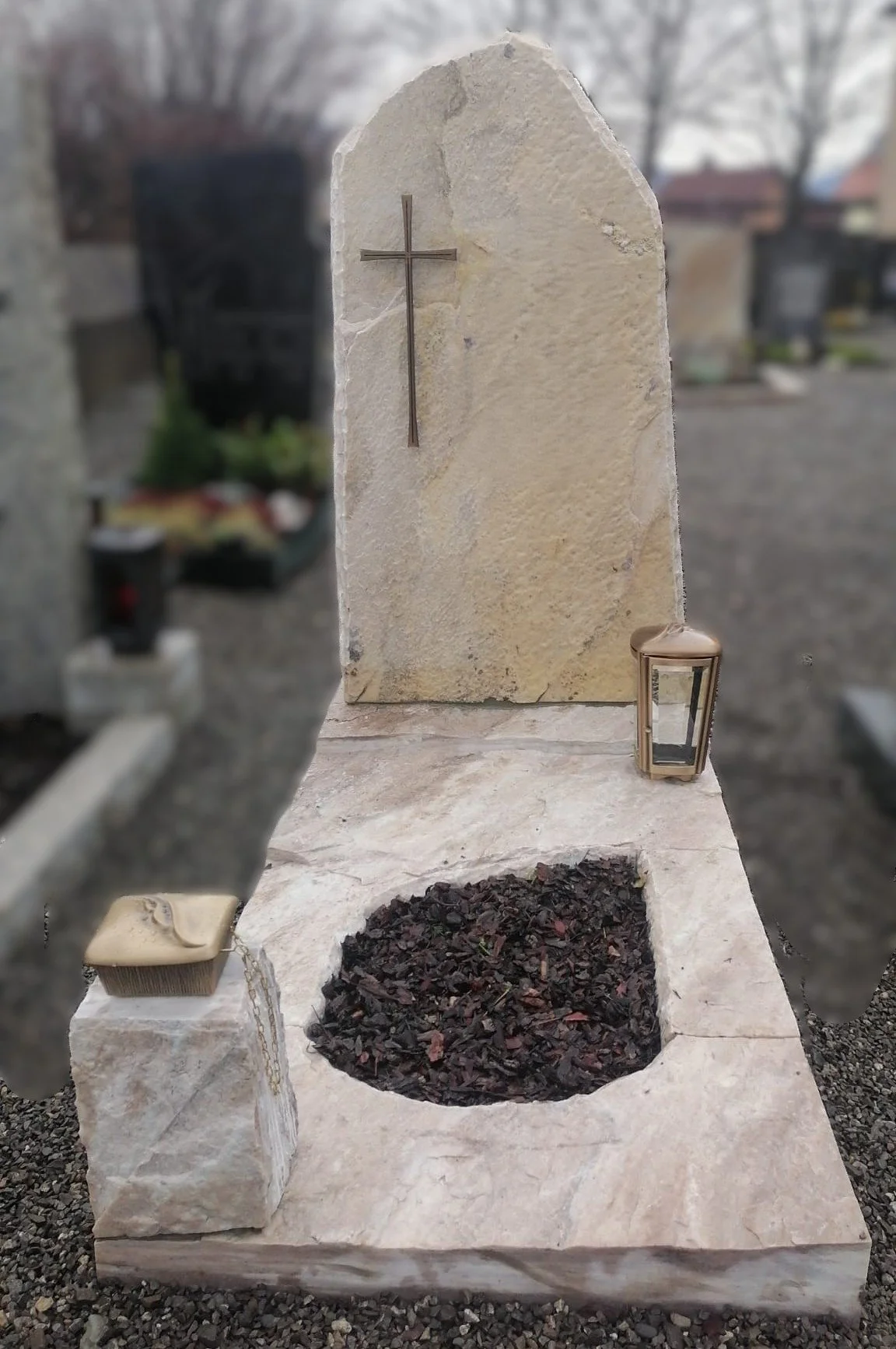 Gedenkstein mit Metallkreuz, Kerzenhalter und Erde auf einem Friedhof.