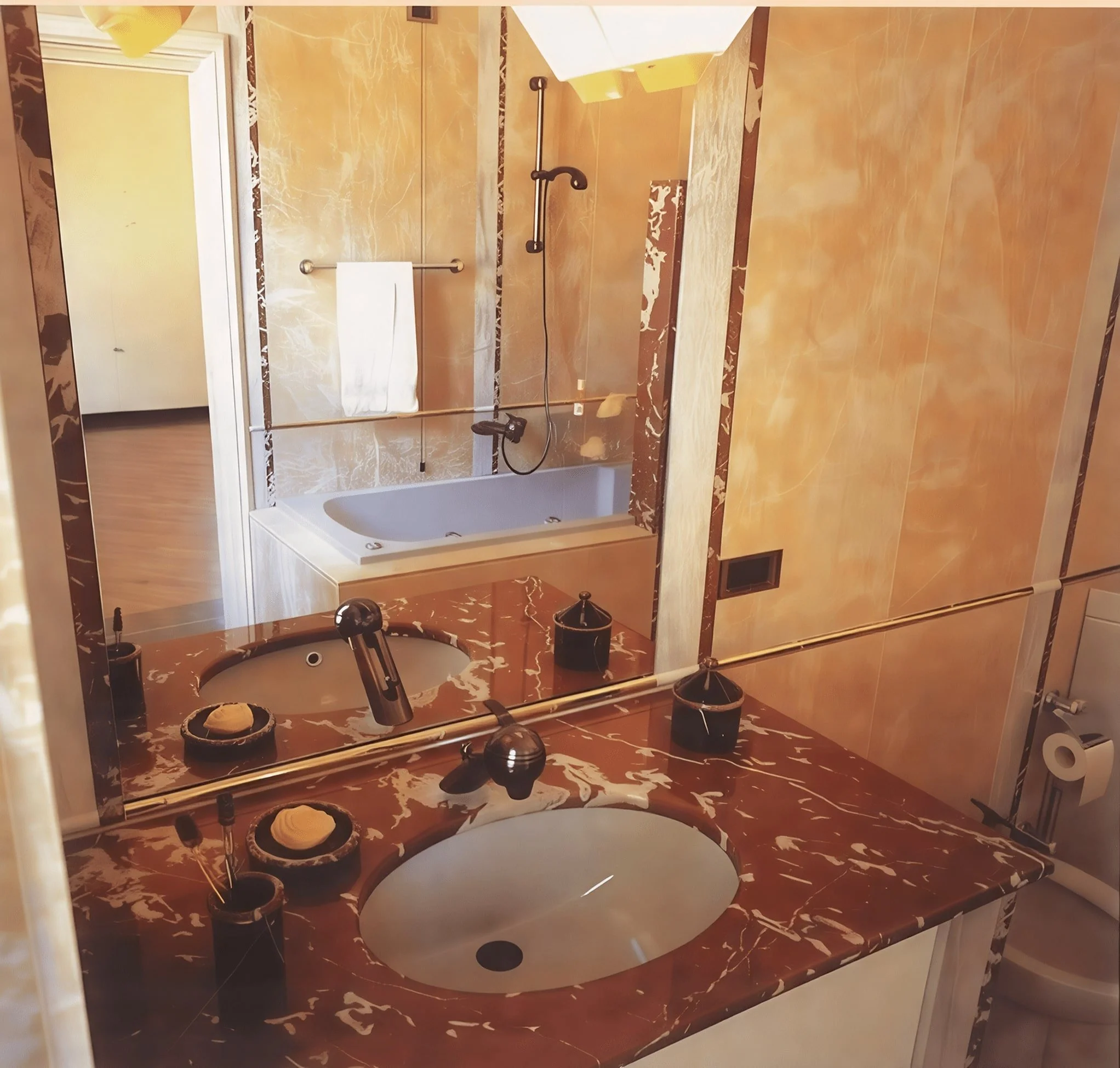 Elegantes Badezimmer mit rotem Marmor-Waschtisch, Doppelwaschbecken, dunklen Handtüchern, Badewanne mit Duschkopf, großer Spiegel, gelben Wänden und Toilette im Hintergrund.