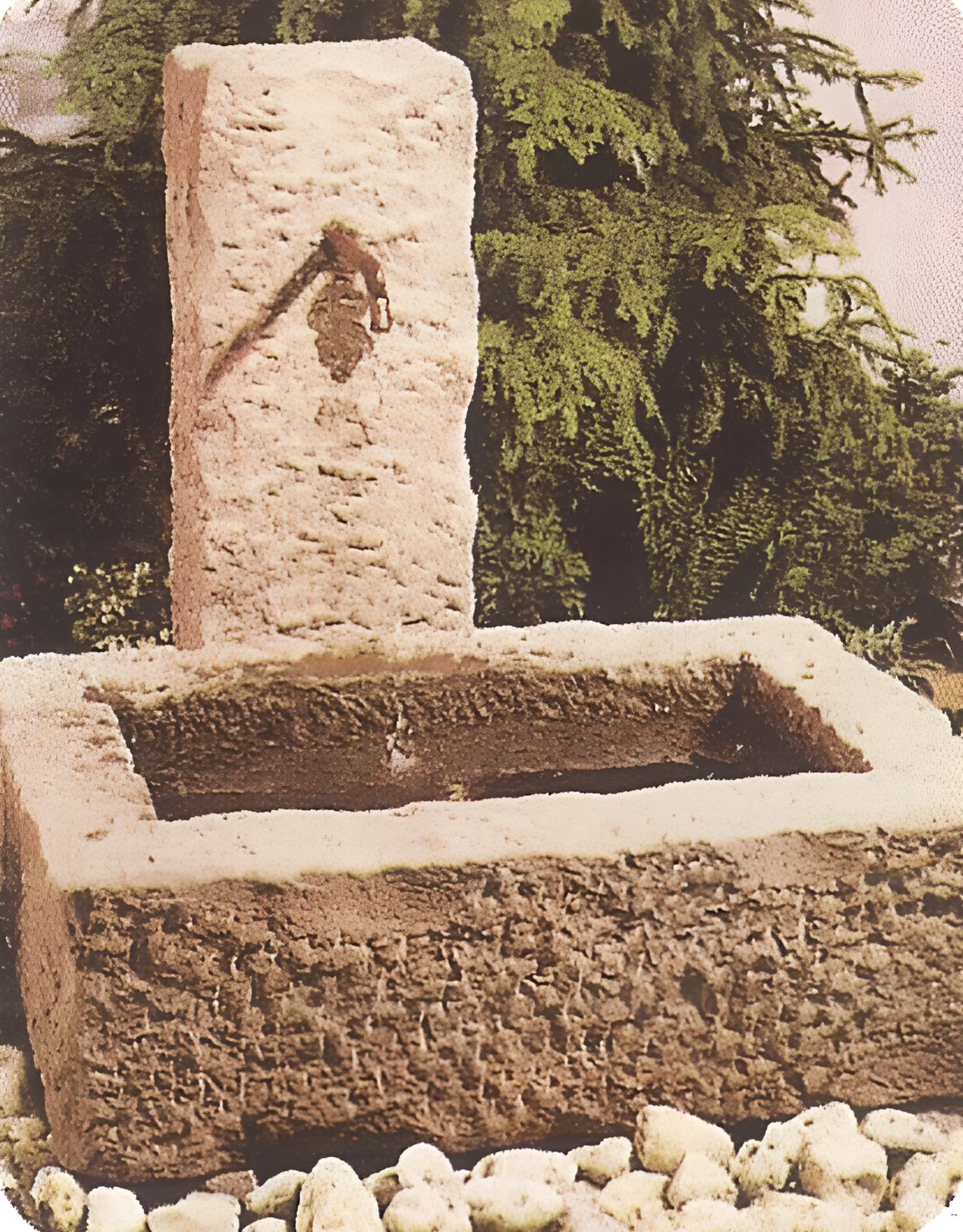 Ein alter Steinbrunnen mit einer Metallfigur eines Künstlers, der einen Pinsel hält, in einem Garten mit grünen Büschen im Hintergrund. Der Brunnen ist quadratisch mit einer hohen Säule.
