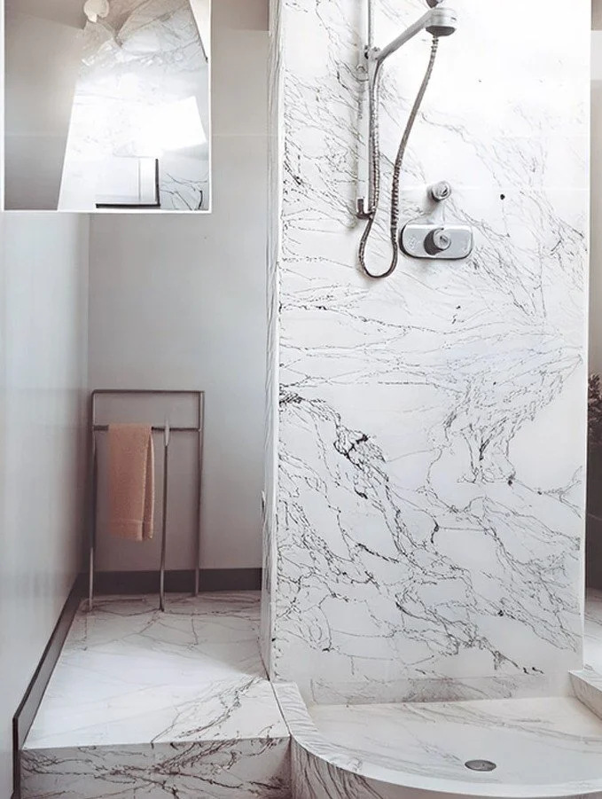 Duschbereich mit weißem Marmor mit schwarzen Adern, Duschkopf und Wassersteuerung, Handtuchhalter mit Handtuch, kleine Fenster oben.