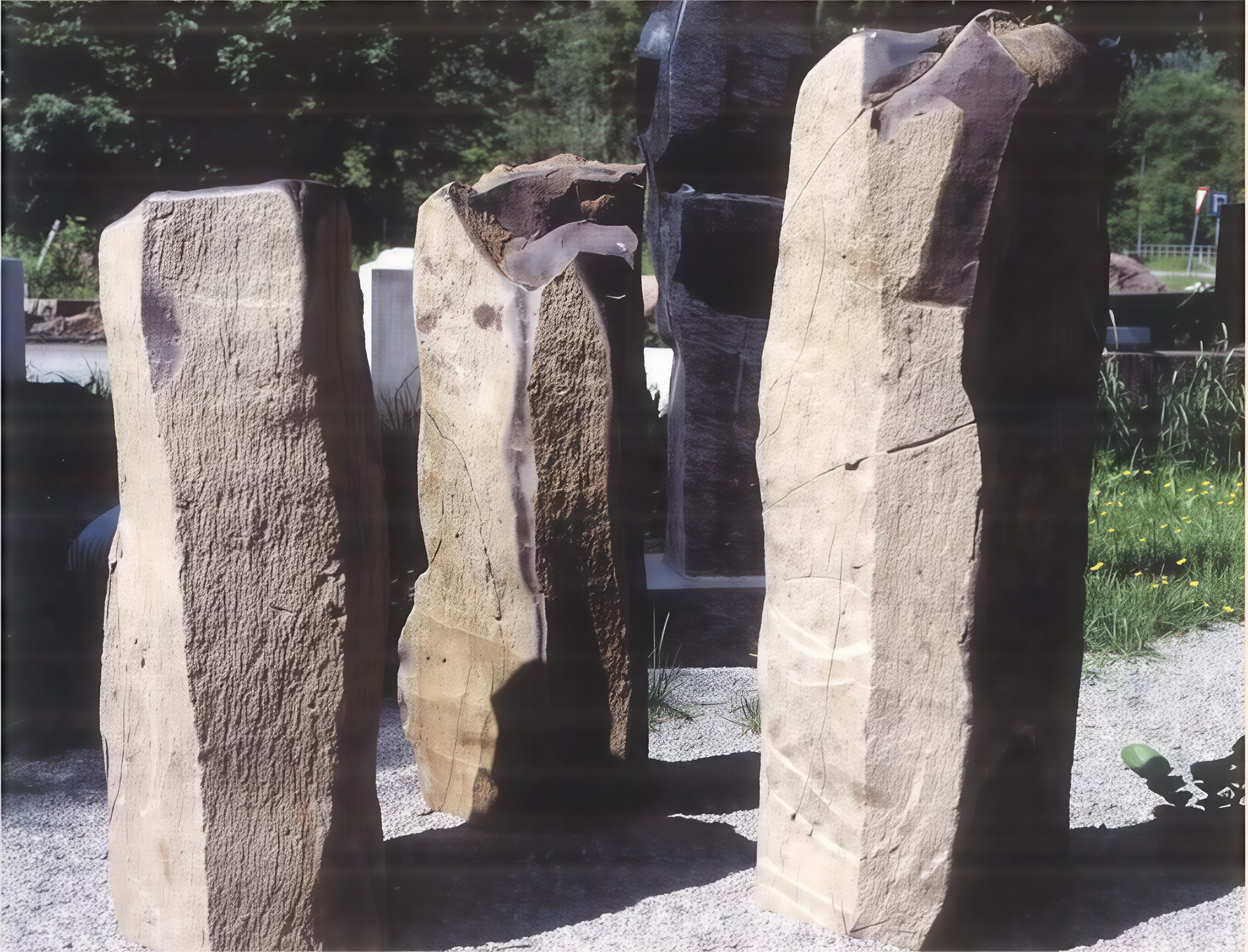 Große Steinskulptur aus mehreren Steinen auf gepflastertem Boden im Freien, mit Bäumen im Hintergrund.