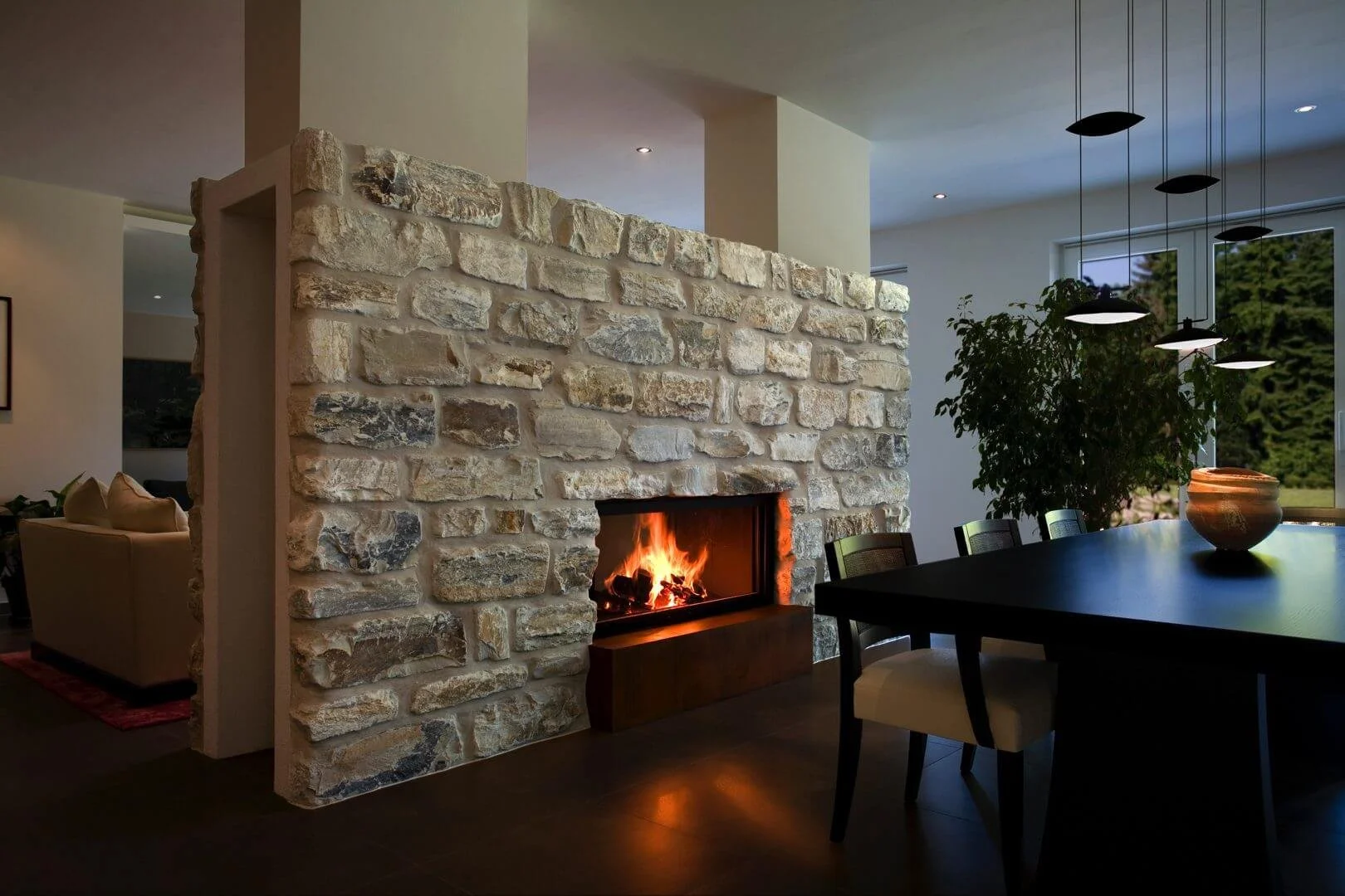Innenaufnahme eines Wohnzimmers mit einem offenen Kamin aus Stein, der ein Feuer entfacht hat, und einem Esstisch mit Stühlen, einem großen Fenster und einer grünen Pflanze.
