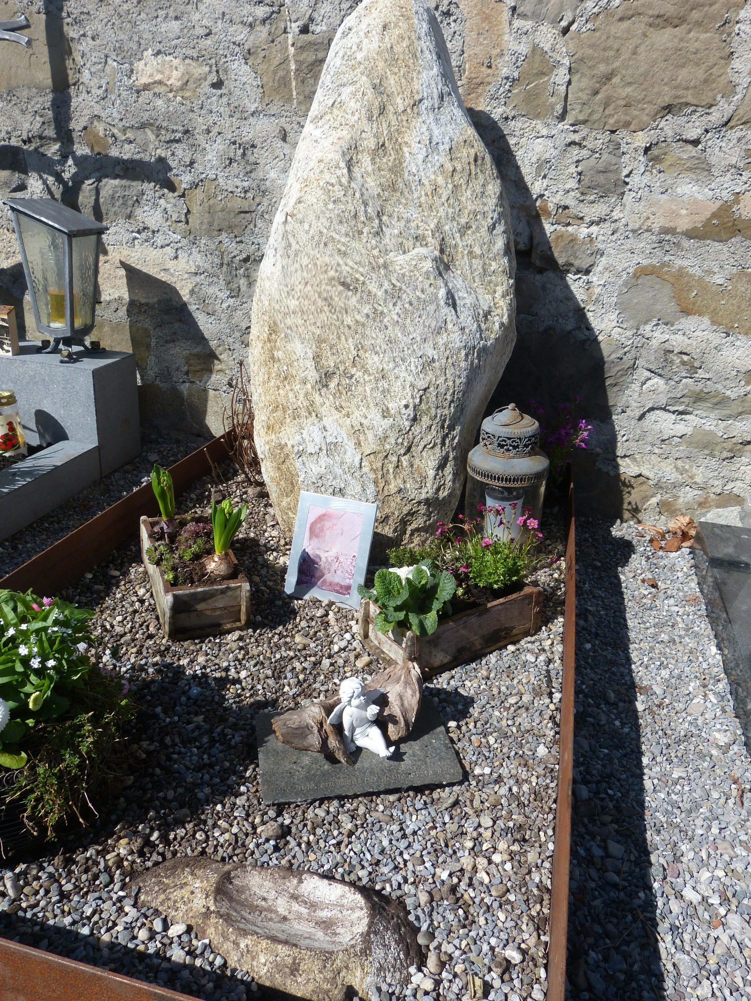 Grab mit Blumen, Kerzen, einem Stein mit Christusfigur und einem großen Findling vor einer Steinmauer