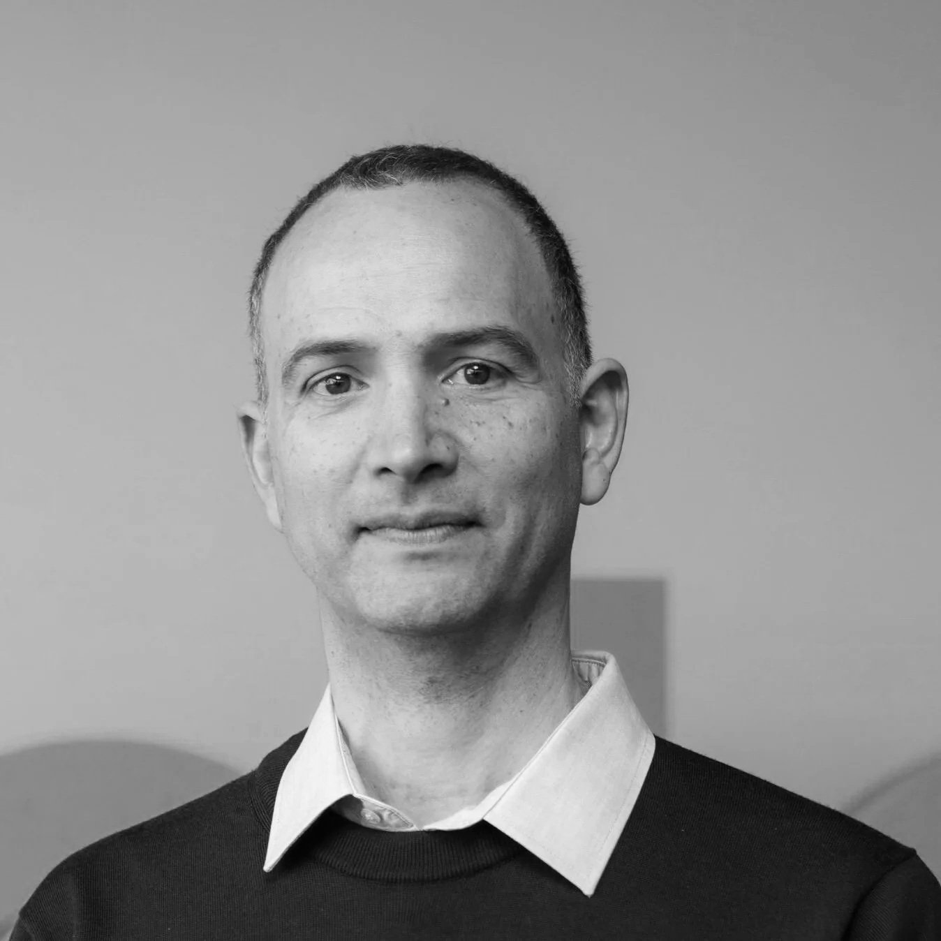 Black and white portrait photo of a man with short hair, wearing a collared shirt and sweater, looking at the camera with a neutral expression.
