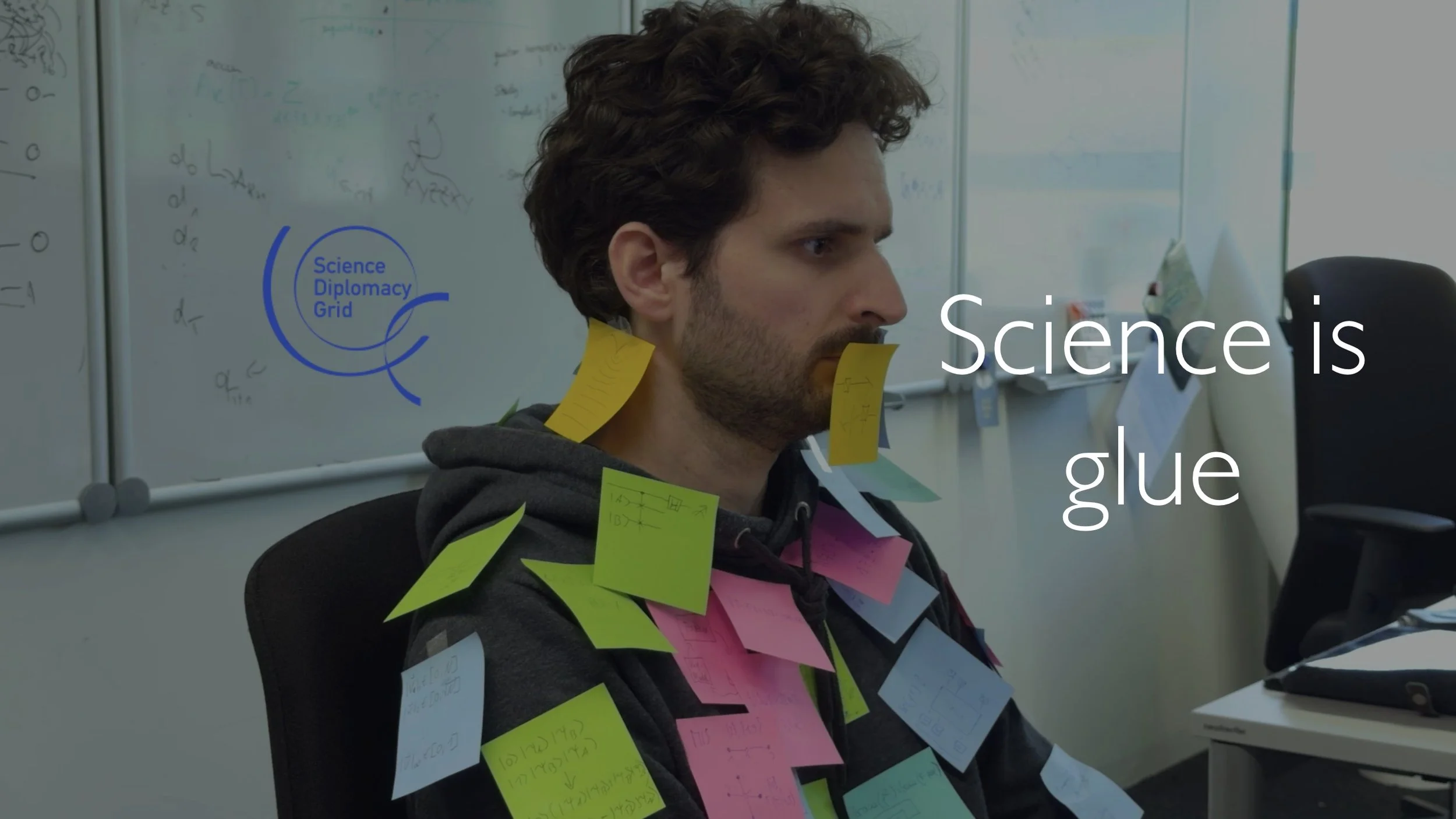A man sitting in an office covered with colorful sticky notes on his shoulders, chest, and face. A whiteboard with writing is in the background. The text on the image reads 'Science is glue'.