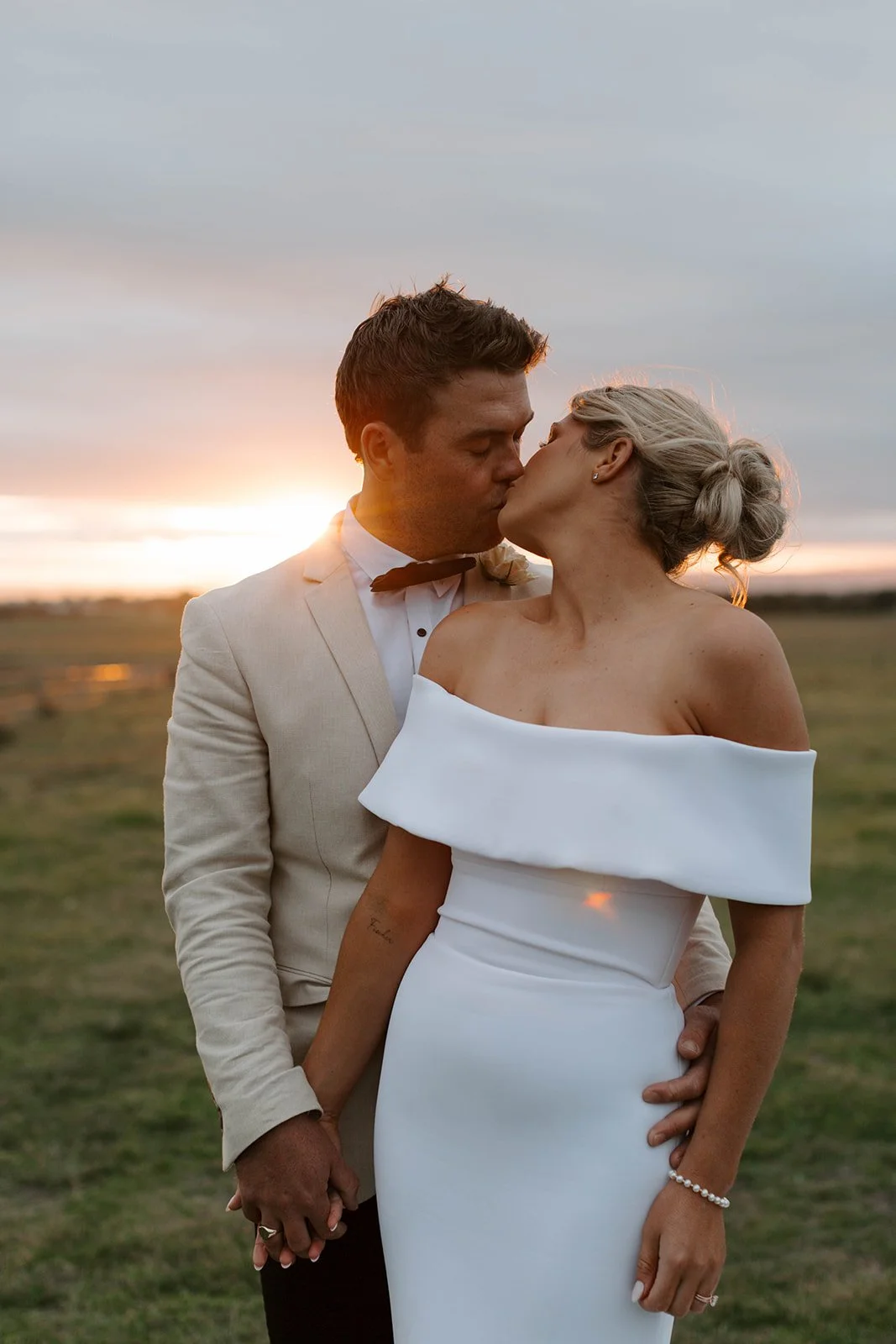 A newlywed couple sharing a kiss during sunset in an open field.