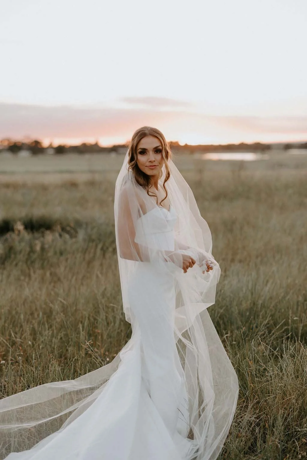 A bride in a white wedding gown and veil standing in an open field during sunset, looking at the camera.