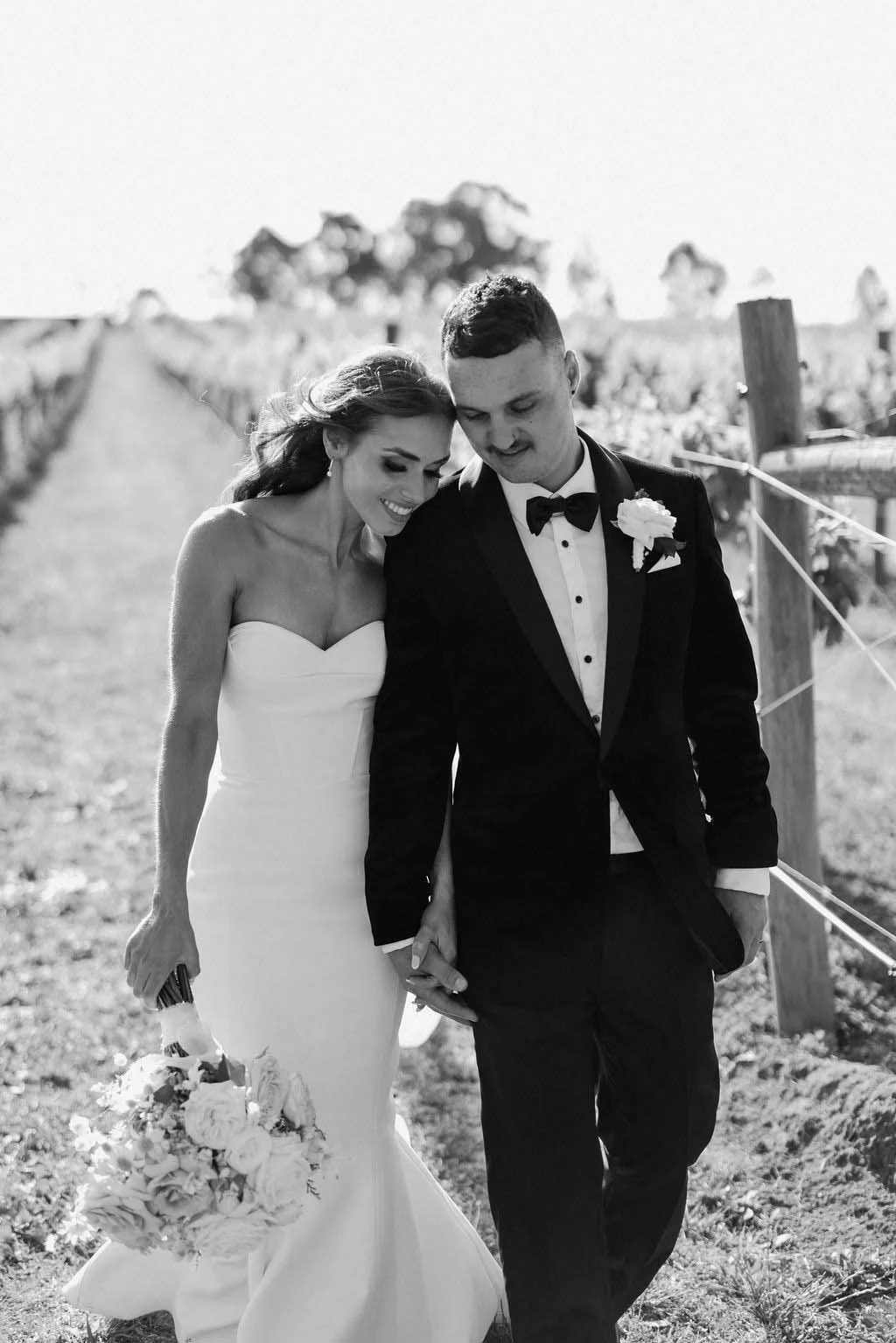 Black and white photo of a bride and groom walking together outdoors, smiling and holding hands, with the bride carrying a bouquet of flowers.